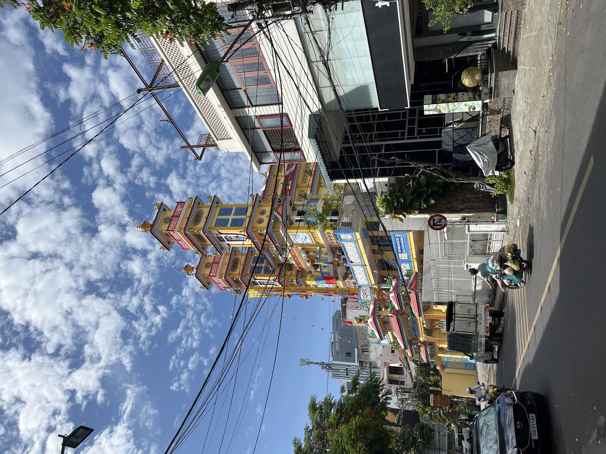 The image shows a street scene in Nha Trang, Vietnam, with a prominent colorful building featuring multiple tiered roofs and towers in the background. The building appears to be a temple or pagoda, decorated with bright yellow, red, and blue accents, and adorned with flags. In the foreground, a dark-colored car with license plate "79A-511.83" is parked on the left side of the road. A motorbike with a rider wearing a white shirt and helmet is seen in front of the car, heading away from the camera. Further down the street, a small truck with a covered bed is parked. Another motorbike, carrying a significant load of goods covered in plastic bags and cloth, is being ridden by a person wearing a gray shirt and helmet, moving from left to right. The street is paved with asphalt and has yellow lane markings. Utility poles with a dense network of electrical wires are visible across the sky. The sky is blue with scattered white clouds. Trees with green foliage and some orange flowers are present on the right side of the street. The building on the right side of the street has a modern architectural style with large windows and a black awning. There are also signs and a large golden spherical object displayed outside this building. The scene captures a slice of urban life with traffic, architecture, and infrastructure typical of a Southeast Asian city.