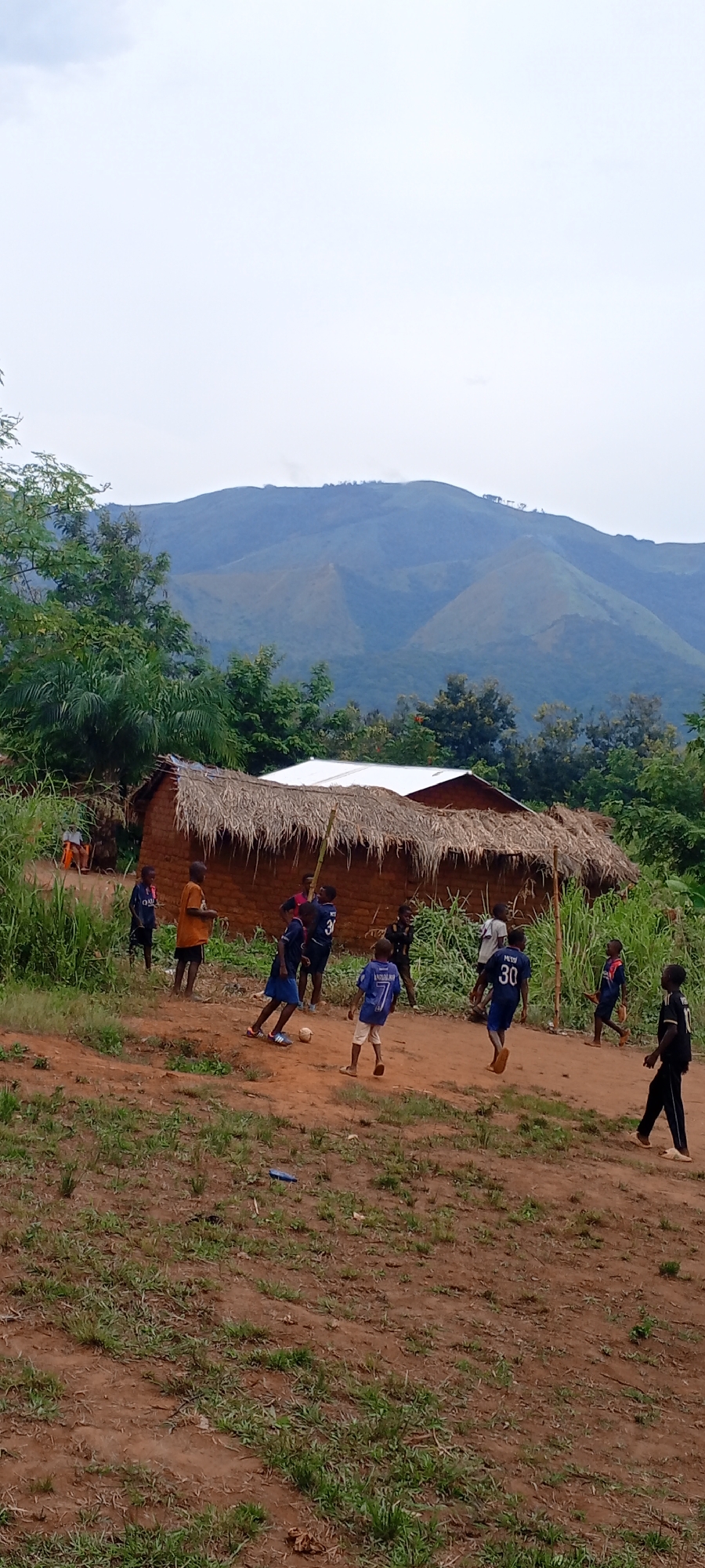 This media file captures a vibrant outdoor scene in Ngalula, Democratic Republic of the Congo, centered around children playing soccer.
In the foreground, a group of approximately eight to ten children are engaged in a game of soccer on a reddish-brown dirt clearing interspersed with patches of green grass. A light-colored soccer ball is visible near the center of the playing area. Many children are barefoot or wearing sandals, and some are dressed in soccer jerseys. Visible text on these jerseys includes "MESSI 30" on at least two shirts, "QATAR" on another, and "ANDALUSIA 7" on a blue jersey. Their body language suggests active play and enjoyment.
In the midground, several simple, traditional houses are visible. These dwellings are constructed from mud bricks with distinctive thatched roofs made of dried grass. At least one house also features a section with a corrugated metal roof. Lush green vegetation, including tall grass, bushes, and trees, surrounds the houses and borders the playing area. Another child can be seen seated in the background near one of the houses.
The background is dominated by a majestic landscape of undulating, verdant mountains, indicating the region's hilly or mountainous terrain. The sky above is light gray and overcast, suggesting a diffused light and possibly a cloudy day, rather than direct sunlight. The overall atmosphere appears natural and rural, reflecting daily life in the village. A small blue object, possibly a piece of plastic, lies on the dirt ground to the left.