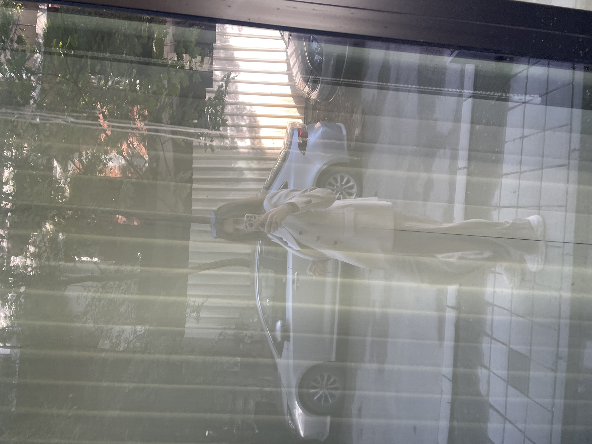 The image, rotated 90 degrees clockwise, presents a scene viewed through a window with horizontal blinds, featuring both reflections and elements beyond the glass. A person, appearing as a reflection, stands prominently in the center, holding a smartphone up to their face, seemingly taking a selfie or recording. They are dressed in a light-colored suit or professional attire, including a jacket and trousers.
Beyond the window and intermingled with the reflection, an outdoor environment is visible. Two light-colored cars are parked, one more fully visible than the other, suggesting a street or parking area. Lush green trees and foliage are present in the background, implying a natural or suburban setting. A striped structure, possibly a fence or part of a building, can also be seen. The bright illumination indicates it is daytime with strong sunlight, contributing to the prominent reflections on the window. No discernible text is visible anywhere in the image.
