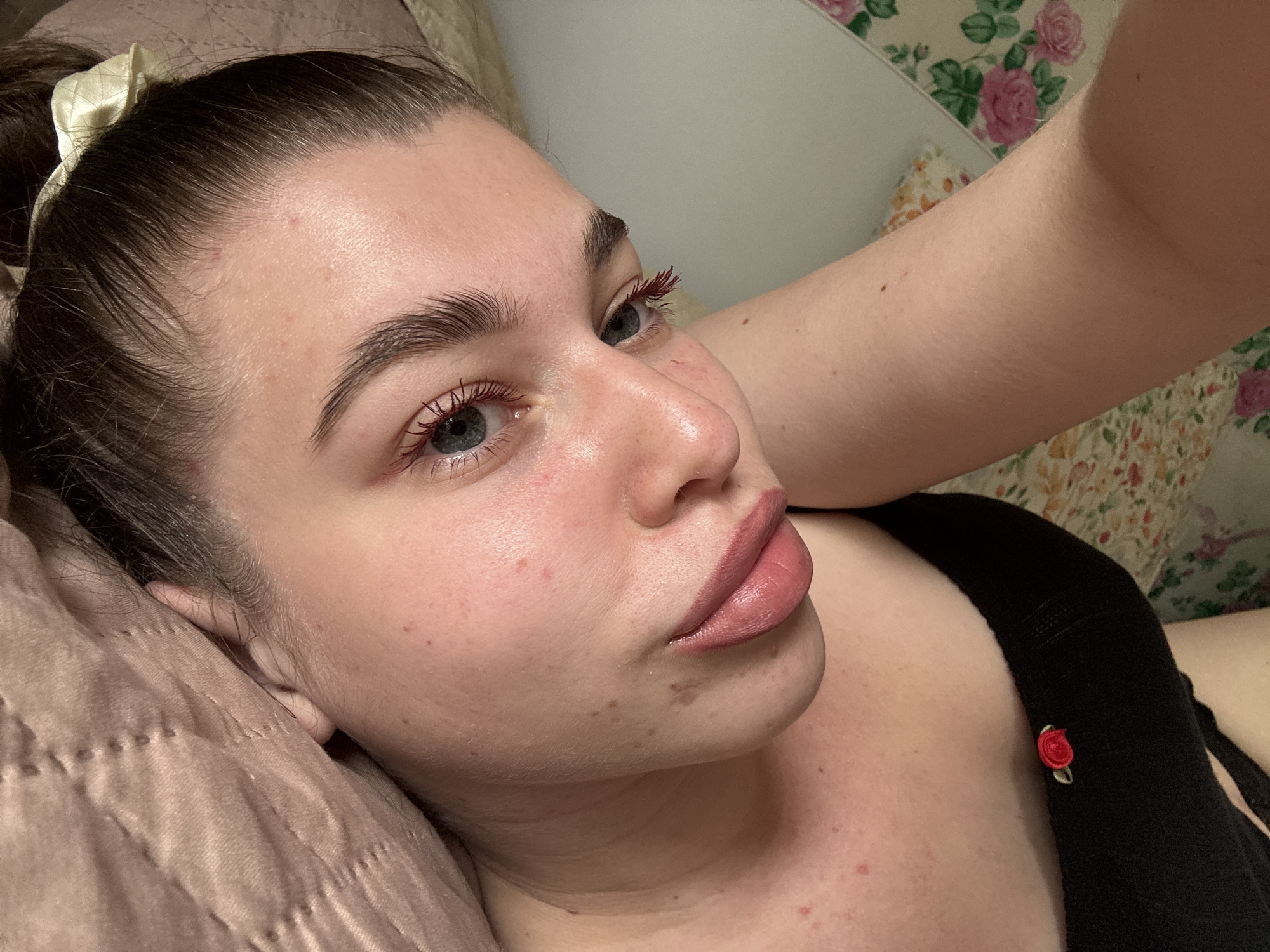 A close-up, angled portrait of a fair-skinned person with light blue eyes. The person is lying on a light brown pillow. The person has dark hair pulled back with a light-colored scrunchie. They are wearing a black top with a small red rose embellishment. The person is wearing red mascara. The background includes a floral patterned surface. Location is unspecified.