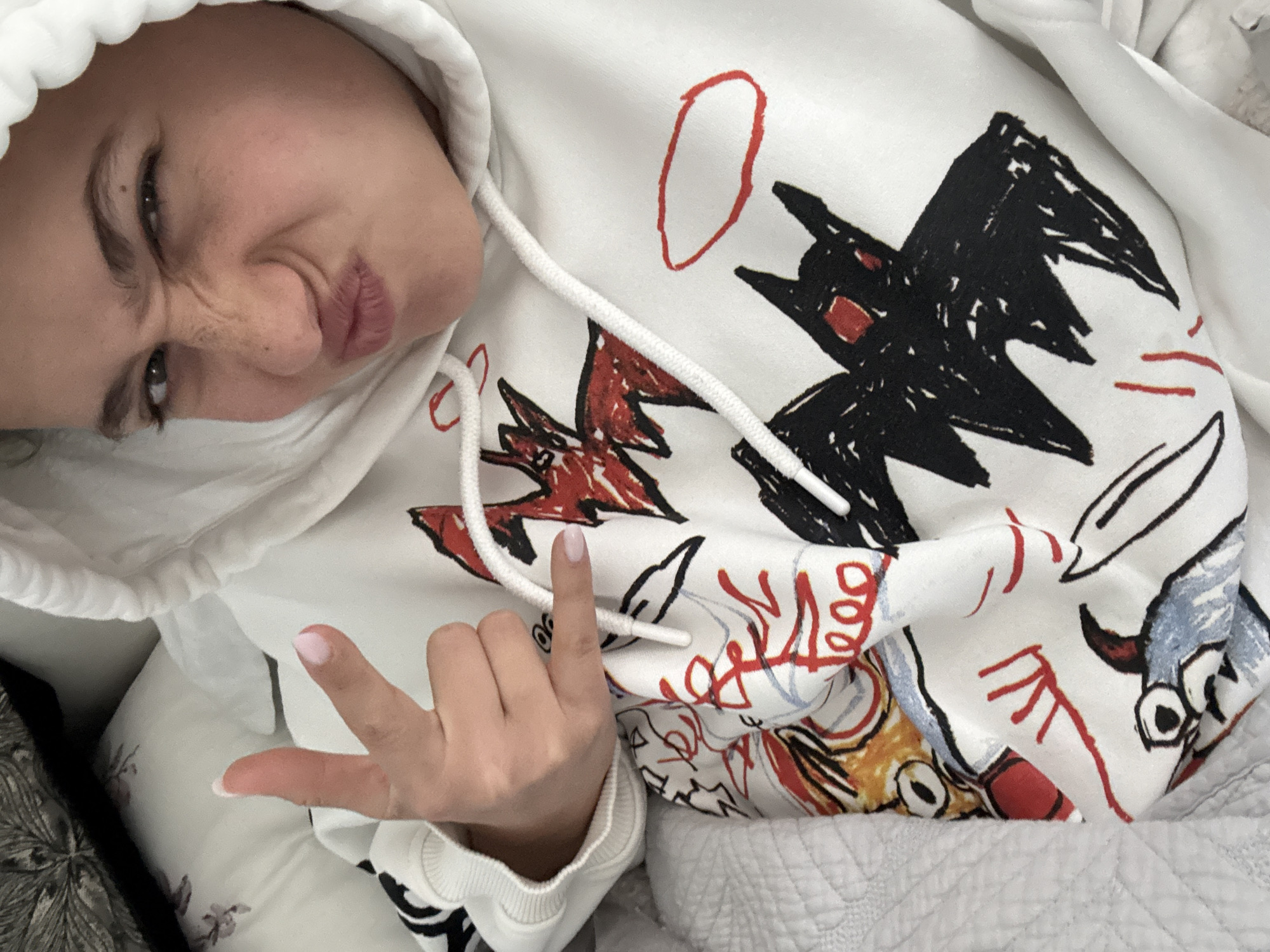 A person is photographed from a high angle, wearing a white hoodie with colorful graphic designs. The person is making a face with pursed lips and a wrinkled nose, and their left hand is making a "rock on" gesture. The background includes a white quilted surface, possibly bedding, and a dark, patterned object on the left edge, potentially a pillow or furniture. There is no visible information to indicate this photo was taken in Milan, Italy.
