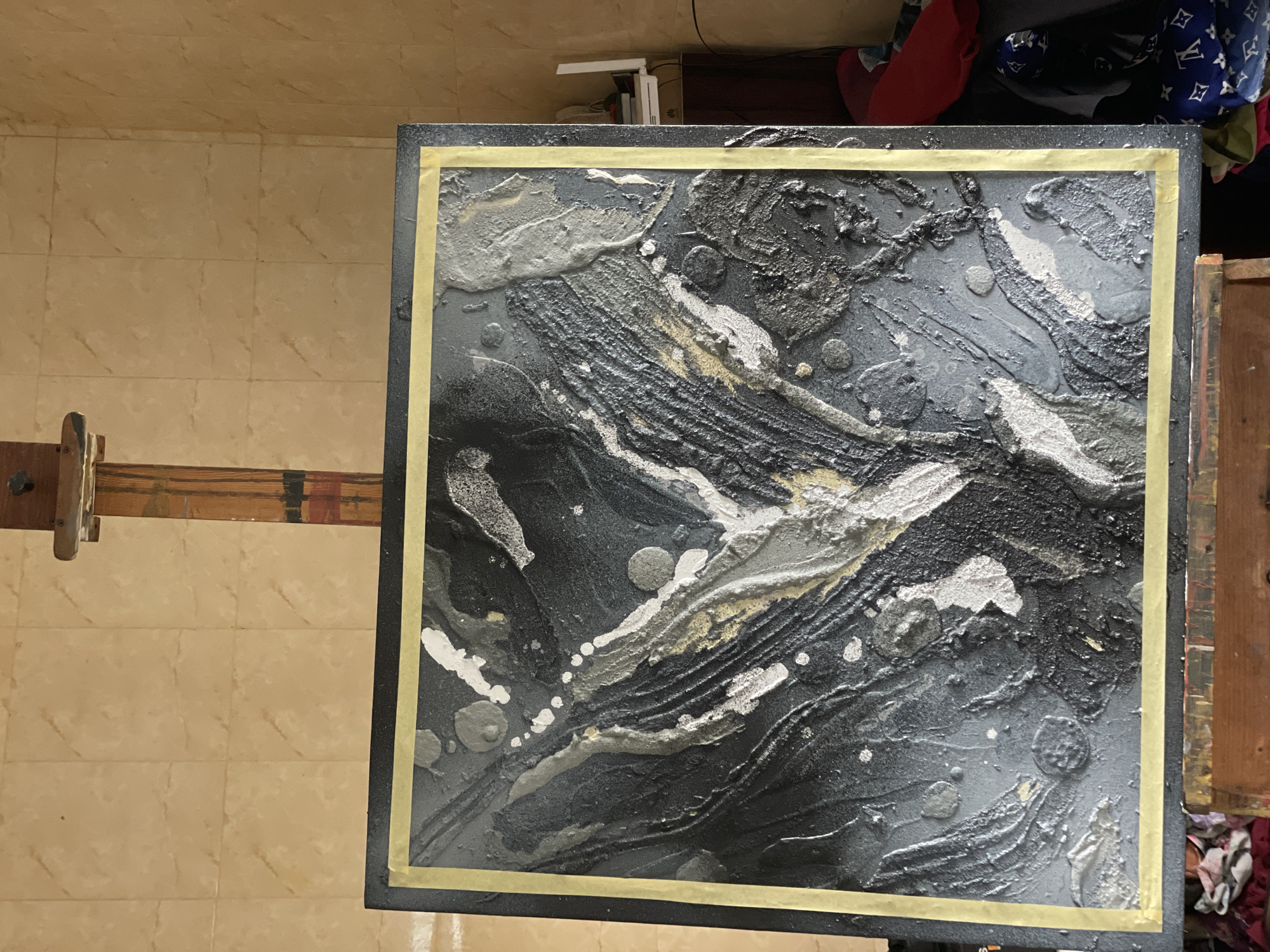The image shows a square abstract painting on an easel. The painting is primarily black, gray, and silver, with textured surfaces and some gold accents. It is framed by yellow tape. The easel is wooden and appears to be adjustable. The background includes a tiled floor, a cabinet, and some clothing. The painting is likely located indoors but cannot be pinpointed to Ado, Nigeria with the information available. There are no people in the image. The primary subject is the abstract painting.