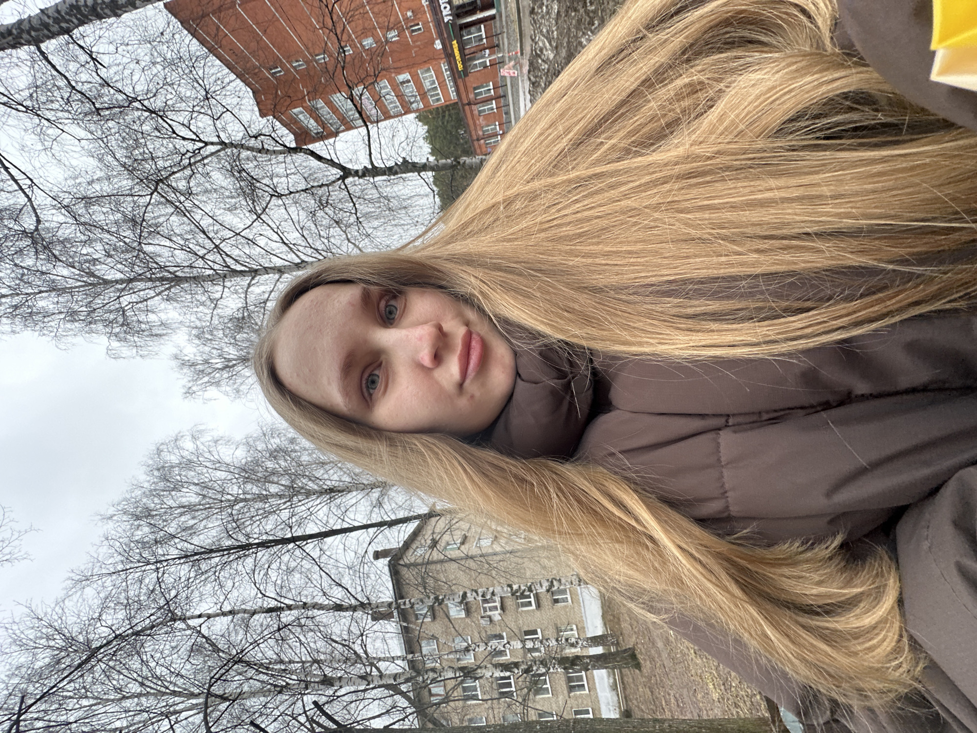 A young woman with long blonde hair is the primary subject, presumably taking a selfie. She is wearing a brown puffer jacket. The background reveals a city environment, with buildings partially visible, suggesting an urban area. Leafless trees are scattered throughout the scene, implying a colder season, potentially late fall or winter. The image appears to have been taken in Chaikovsky, Russia, based on the architectural style of the buildings and environmental characteristics.