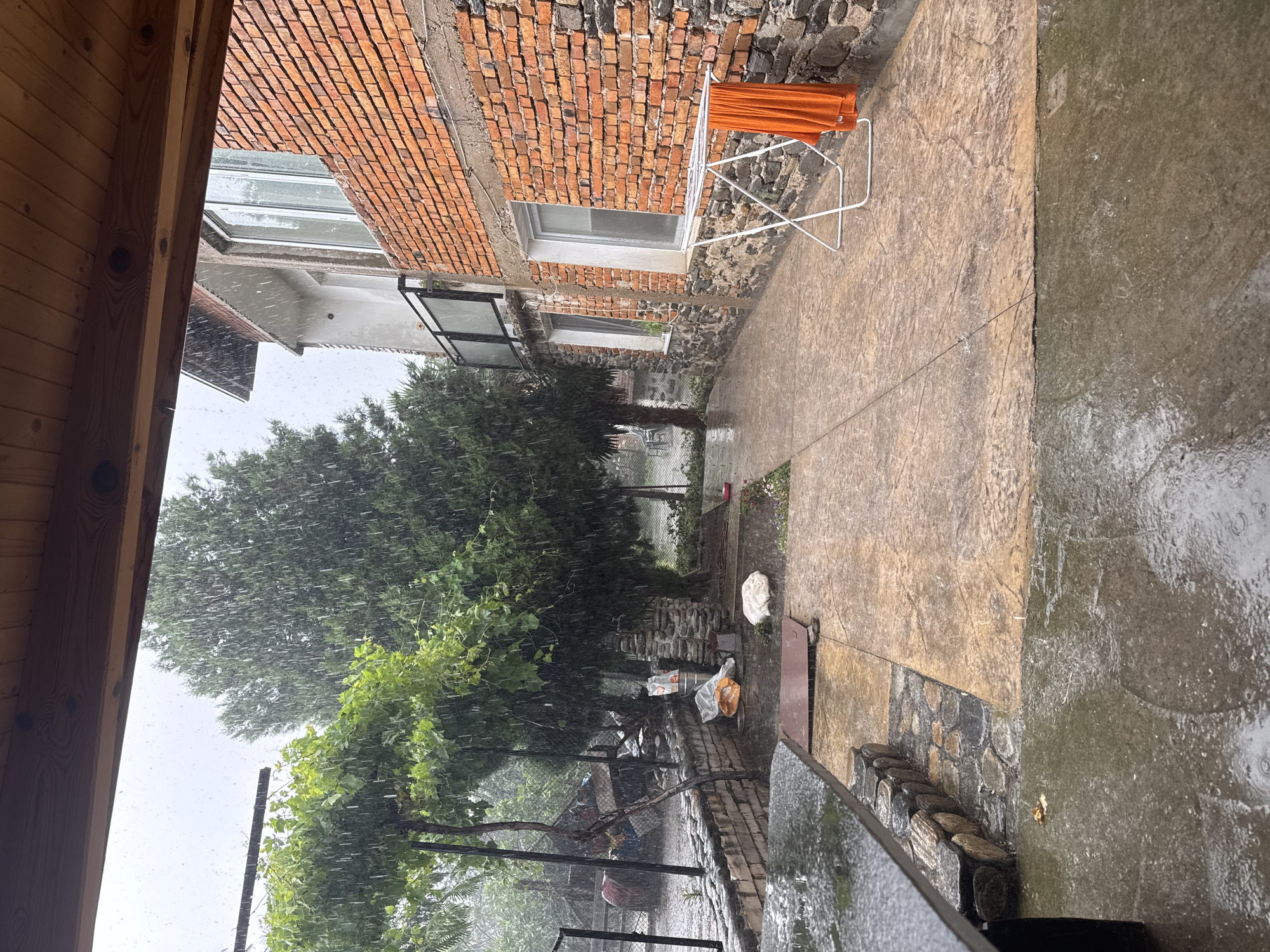 The image depicts an outdoor scene during a heavy downpour. The foreground shows a wet, dark stone surface of what appears to be an outdoor table or bench, with water pooling on it. Beyond this, a patterned concrete patio is also wet and has puddles. To the right of the patio is a white drying rack with a vibrant orange towel hanging on it. A brick building with stone foundations occupies the right side of the image, with windows visible. Trees with lush green foliage are present in the background, also being heavily rained upon. The sky is overcast and grey, indicating continuous rainfall. The scene appears to be a residential outdoor space, possibly a backyard or patio area. Based on the provided context, this scene is located in Parvenets, Bulgaria.