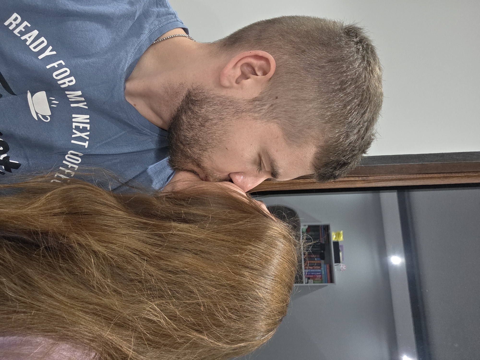 A man and a woman are kissing indoors. The man has short brown hair, a beard, and is wearing a blue t-shirt with the text "READY FOR MY NEXT COFFEE." The woman has long brown hair. A reflection of a bookshelf is visible in the background. There is no information to suggest that this image was taken in the city of Yagodovo, Bulgaria.