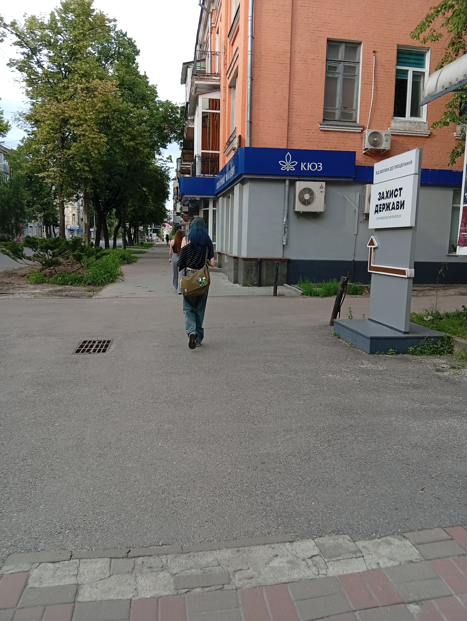 This image depicts a street scene in Poltava, Ukraine. The view is from a low perspective, looking down a street with a paved road on the left and a tiled sidewalk on the right. In the foreground and mid-ground, a person with blue hair is walking away from the camera. This individual is wearing a dark and light striped, possibly off-shoulder, short-sleeved top, blue jeans, and dark shoes with white soles. They are carrying an olive green messenger bag adorned with multiple circular pins or badges on its flap. Further down the sidewalk, a second person, less distinctly visible, is also walking away. To the right, a multi-story building features a reddish-orange brick facade on its upper section and a light grey facade on its lower section. Two windows are visible on the upper brick wall: an older-style multi-pane window and a more modern, white-framed window with a partial blue and white blind. Below the modern window, a grey outdoor air conditioning unit with a "CH" logo is mounted. Below the blue sign on the grey facade, another white outdoor air conditioning unit with a "MITSUBISHI" logo is visible. A prominent blue sign with white lettering and a white leaf-like logo displays the text "КІОЗ" (KIOZ). Adjacent to this, a vertical white sign post features text at the top "ВІД ВЕЛИКИ ВІДІВ ДО ПРОЦВІТАННЯ" (FROM GREATNESS TO PROSPERITY), followed by the main text "ЗАХИСТ ДЕРЖАВИ" (DEFENSE OF THE STATE), and smaller text "ГРОМАДСЬКА ОРГАНІЗАЦІЯ" (PUBLIC ORGANIZATION). An L-shaped brown arrow points left and then upwards on this sign. The sign post is anchored in a low, dark grey concrete base, surrounded by sparse green vegetation. A lean wooden post rests against the building's grey base nearby. The asphalt road on the left is dark grey and granular. A square metal storm drain grate is embedded in the asphalt near the curb line in the mid-ground. The sidewalk on the right is composed of rectangular pavers in reddish-brown and grey tones, showing signs of wear and minor cracks. A raised concrete curb separates the road from the sidewalk. A narrow strip of soil with small green plants and weeds runs between the sidewalk and the building facade. On the left side of the street, a line of tall, mature trees with dense green foliage forms a canopy, with some leaves exhibiting yellowish hues. Beyond the trees, other buildings are partially visible
