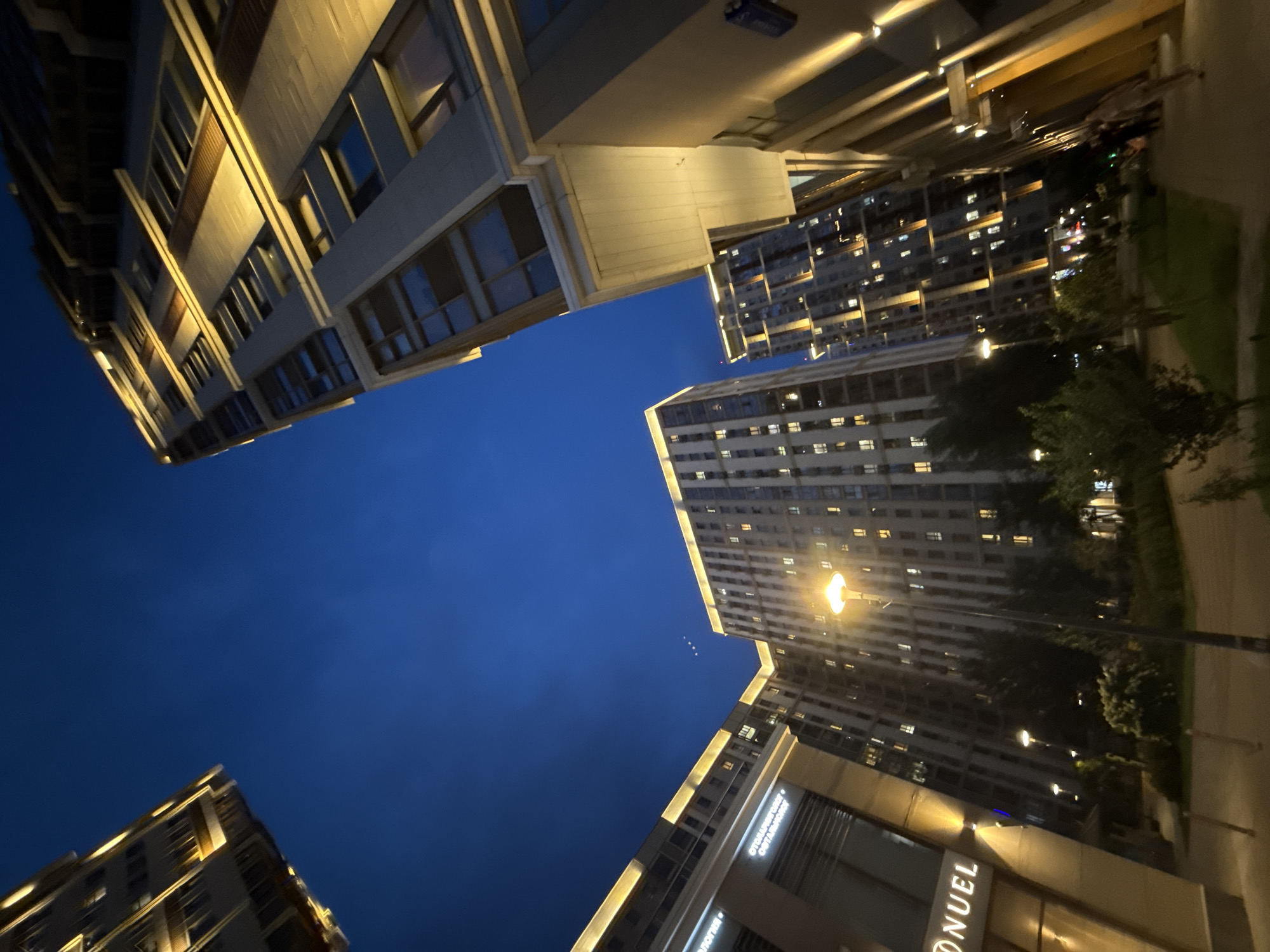 The image depicts a wide-angle, low-angle perspective of modern residential buildings at dusk in Moscow, Russia. The buildings are tall and feature contemporary architectural designs with illuminated facades and numerous windows. The sky is a deep blue, transitioning into twilight. In the foreground, a pathway is visible, with streetlights and some greenery. A few people are walking on the pathway in the distance. The overall impression is one of urban development and evening ambiance. Specifically, this appears to be a view of the residential complex "Dom na Krylatskikh Holmah" in Moscow.