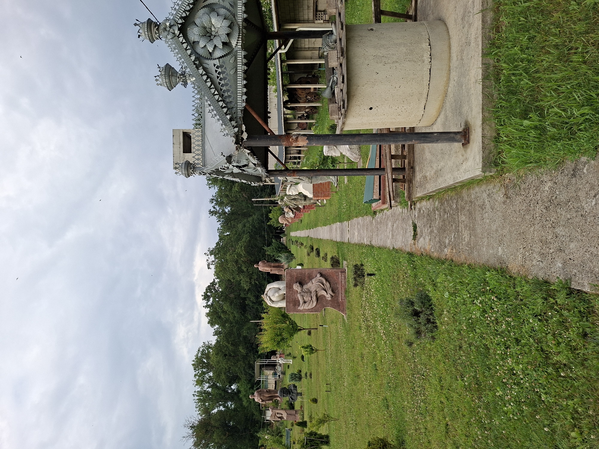 The image depicts an outdoor sculpture park in Malcoci, Moldova. A concrete pathway runs through a grassy area dotted with various statues and busts. On the right side of the pathway, a well structure with a decorative metal roof is prominently featured. The well is made of concrete, and its roof is adorned with intricate metalwork. The park is surrounded by trees and under a cloudy sky. The overall scene conveys a sense of a public space dedicated to art and history.