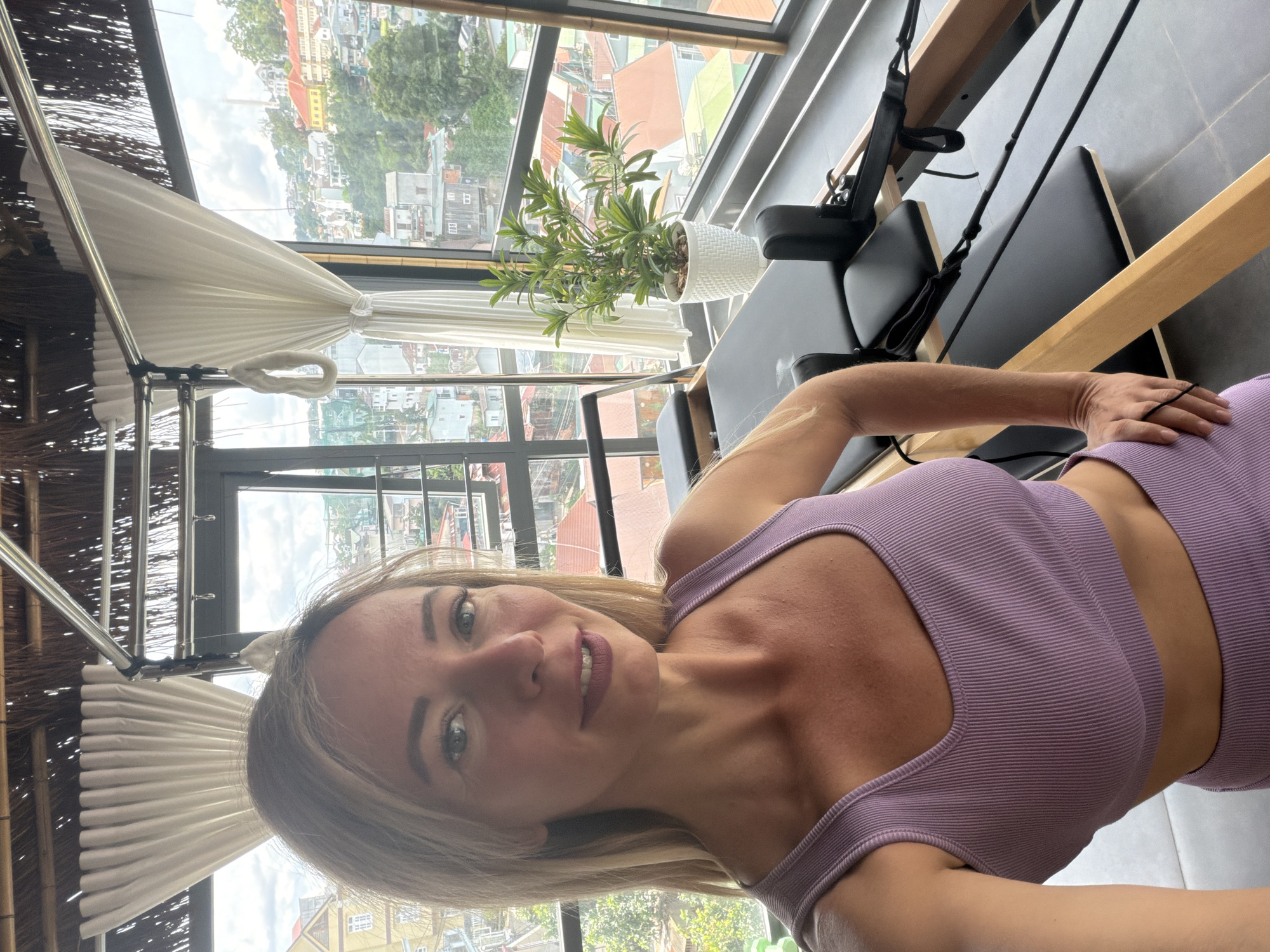 A woman with blonde hair and fair skin, wearing a purple athletic top and matching bottoms, is taking a selfie in a bright room with a view of Da Lat, Vietnam. She is standing to the left of a Pilates reformer. The room features a thatched ceiling, white curtains, and large windows that offer a panoramic view of the hilly cityscape, characterized by colorful buildings and lush greenery. A potted plant sits on the floor near the window. The lighting suggests it is daytime.