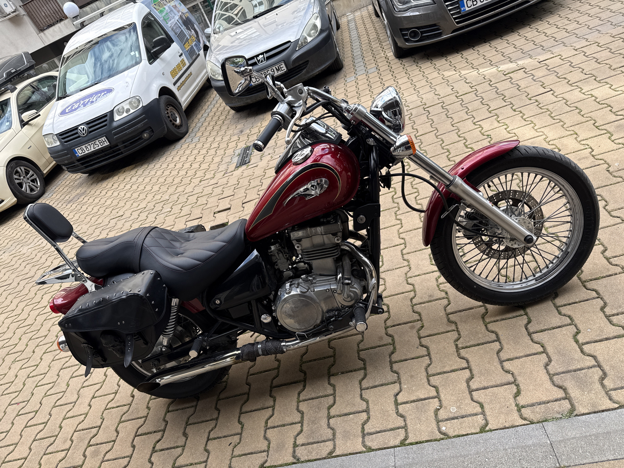 The image displays a stationary outdoor scene in Sofia, Bulgaria, featuring a dark red cruiser motorcycle parked on a light brown interlocking concrete paver surface. The motorcycle is equipped with a dark red fuel tank and fenders, a black textured seat with a passenger backrest, and black leather saddlebags featuring metal studs and a skull emblem. Notable chrome elements include the handlebars, headlight, mirrors, engine components, and exhaust pipes, one of which is partially wrapped in black heat tape. The front wheel is a spoked design with a visible disk brake.
Behind the motorcycle, several vehicles are parked. On the far left, a light-colored sedan is partially visible, fitted with a black roof cargo box. Immediately to its right is a white Volkswagen Caddy van, bearing the Bulgarian license plate CB 8725 BH and displaying a blue "Carrier" logo on its front, along with commercial graphics and a phone number on its side. Adjacent to the van, a silver Peugeot 307 sedan is parked with the Bulgarian license plate CB 7959 ME. On the far right, the front bumper and grille of a dark grey Audi sedan are partially visible, featuring a Bulgarian license plate starting with "CB".
The ground consists of neatly arranged light brown and beige concrete pavers, with some minimal vegetative growth between the blocks. A dark grey concrete curb runs along the lower edge of the image, defining the boundary of the paved area. No individuals are present, and no actions are depicted. The scene is illuminated by natural daylight.