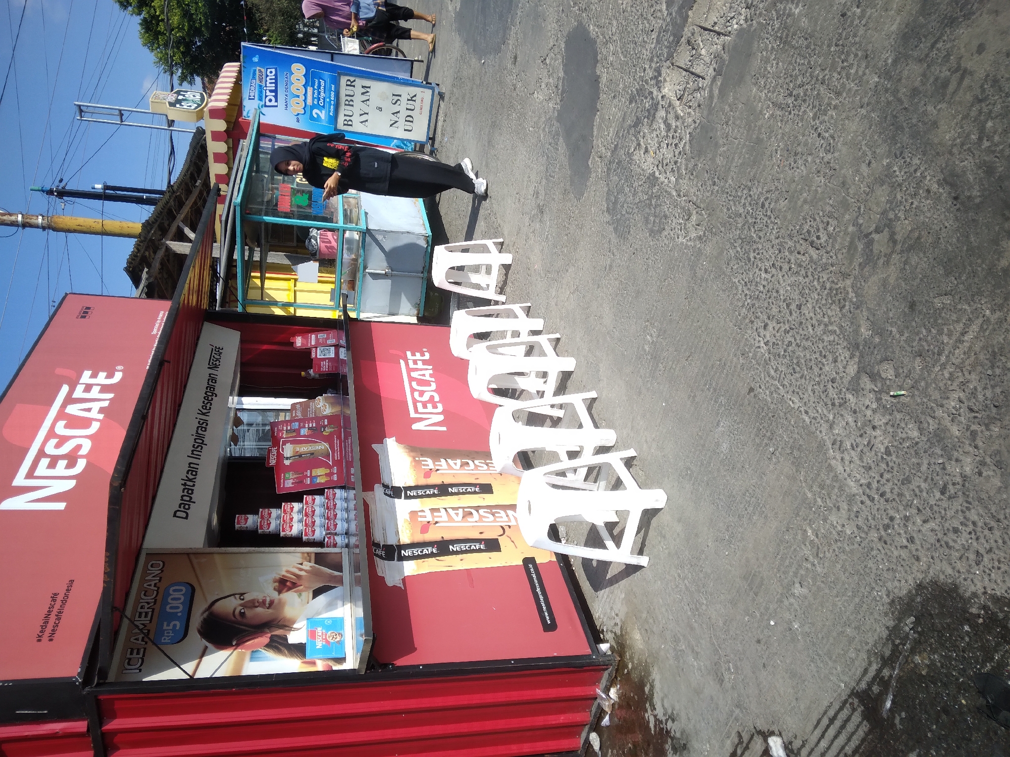 In Purwokerto Kidul, Indonesia, a Nescafé stall is open for business on a sunny day. The stall is a red, corrugated metal structure with a large Nescafé logo prominently displayed. In front of the stall are five white plastic stools arranged in a row on a weathered concrete surface. A woman in a black headscarf and black clothing, carrying a smartphone, is walking past the stall. She appears to be a customer or a passerby. Behind the Nescafé stall, a smaller food stall with a blue and yellow awning is visible. A blue and white sign with Indonesian text, advertising "BUBUR AYAM" (chicken porridge) and "NASI UDUK" (coconut rice), stands near the food stall. The environment is typical of a street-side vendor in Indonesia, with a mix of commercial activity and everyday life. The ground is uneven and shows signs of wear and tear. The sky is clear and blue, suggesting pleasant weather.