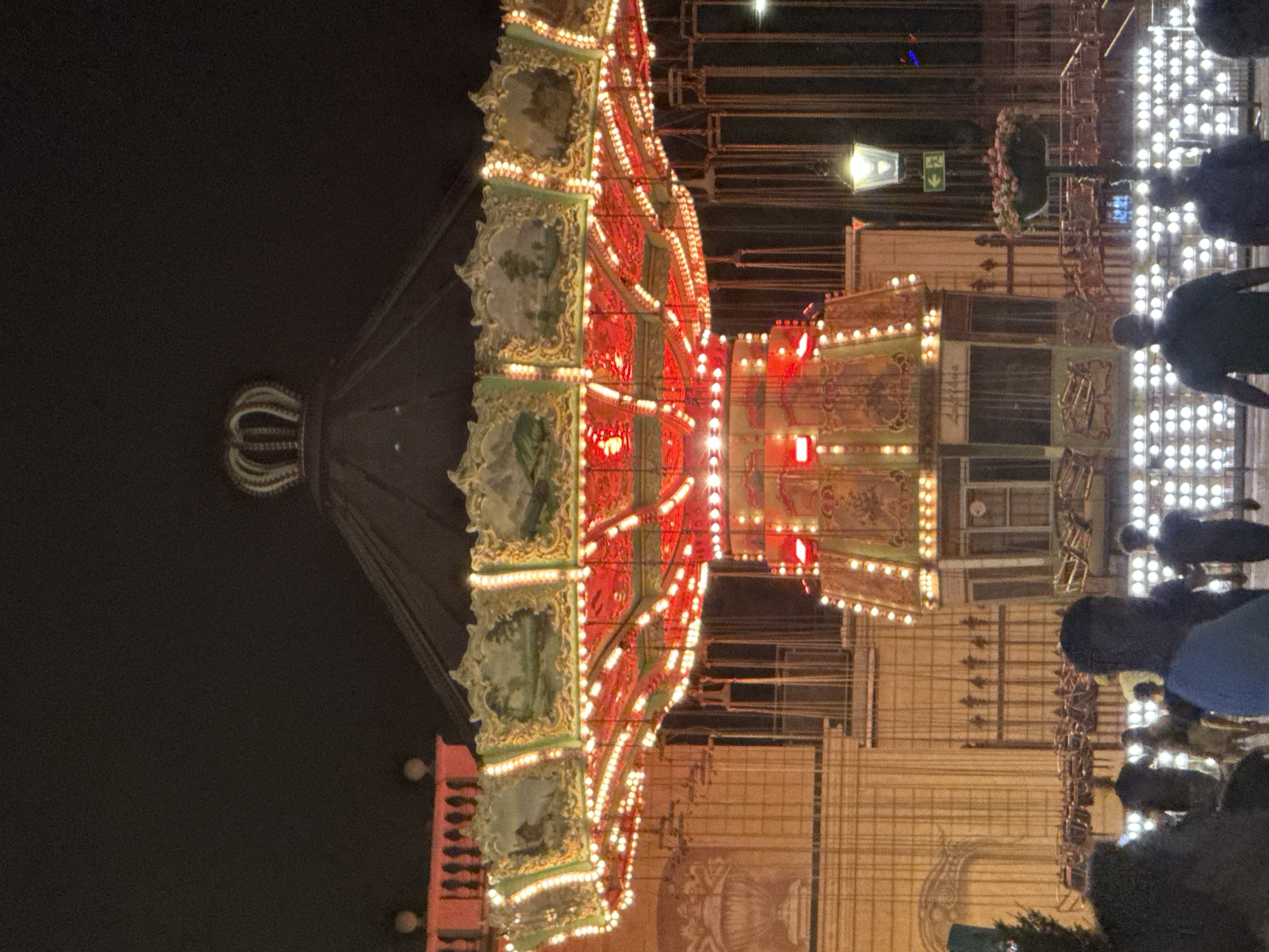 This is a photograph of a swing ride at night, likely at a fair or amusement park. The ride is illuminated with numerous red and yellow lights. The central structure of the ride is decorated with detailed paintings and has a large, ornate crown at the top. In the background, there is a building with architectural details and a streetlamp with a sign indicating an exit. Several people are visible in the foreground and mid-ground, some walking and others standing, appearing to be attending the event. The overall atmosphere is festive due to the nighttime setting and the bright lights of the amusement ride. The context provided by the user indicates the location as Vienna, Austria.