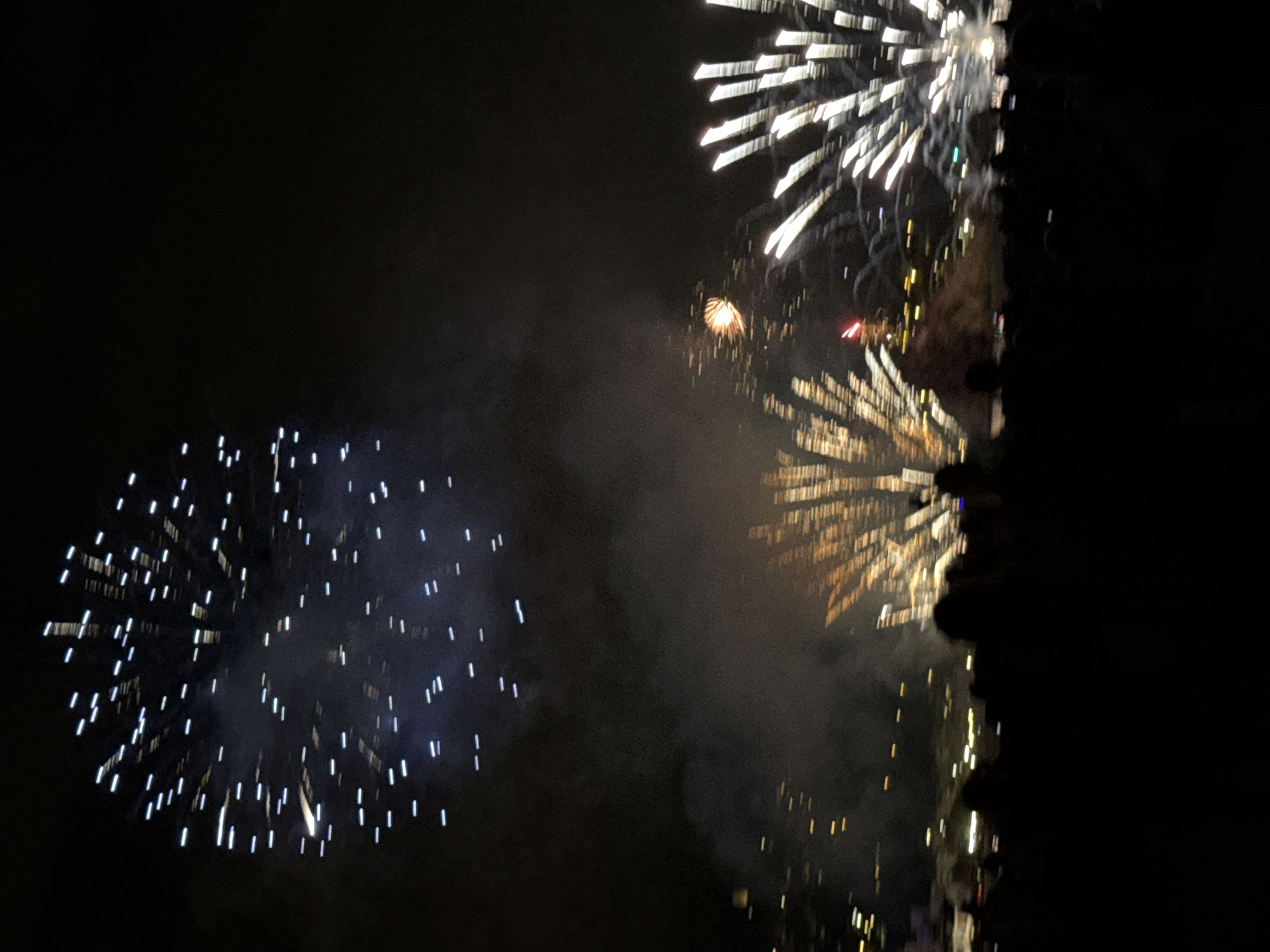 An outdoor fireworks display is underway at night, with a crowd of silhouetted people visible in the foreground. Multiple bursts of fireworks illuminate the dark sky. In the upper left, a large display of white and blue streaks emanates from a central point. In the middle of the frame, a golden firework explodes, with streaks radiating outwards, partially obscured by smoke. To the right, another large firework burst shows white, streaking lights. In the lower portion of the image, the dark silhouettes of many people are gathered, facing the sky. The background shows blurred city lights. The fireworks are consistent with a celebration like the Blanes Festa Major.