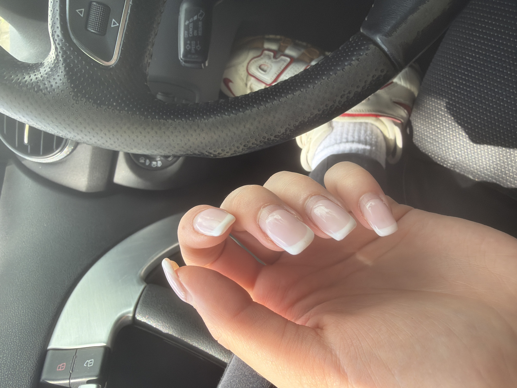 The image depicts a hand with French-manicured nails resting in the interior of a car, specifically on the steering wheel and surrounding dashboard. The steering wheel is black and has various buttons and controls. A partial view of a white sneaker with red accents is visible near the steering wheel. The car seat is black and textured. The scene appears to be inside a vehicle during daytime, judging by the light. There is no evidence to suggest the location is in the city of Smolyan, Bulgaria.