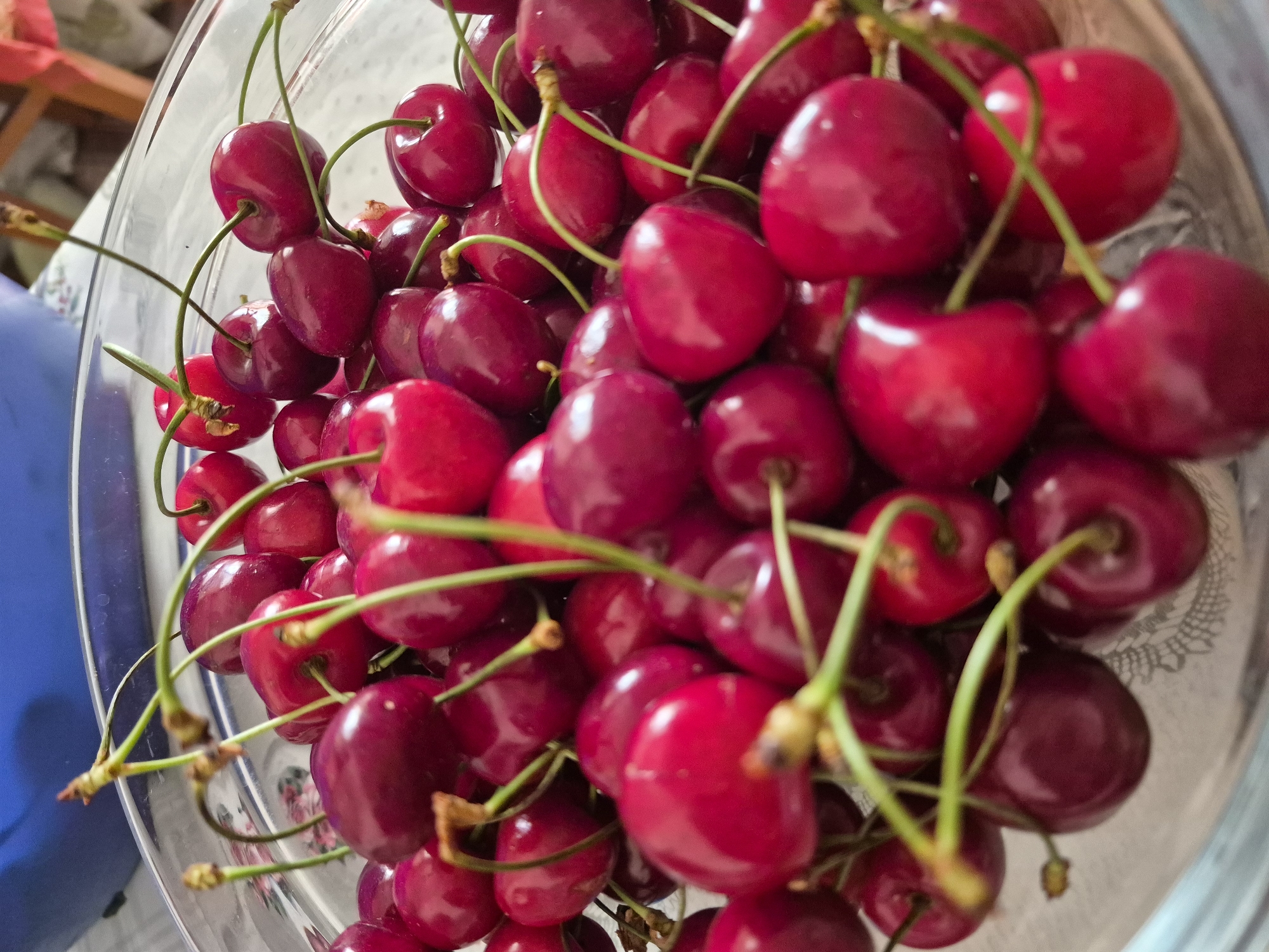 A close-up, overhead view shows a clear glass bowl filled with ripe, dark red cherries. The cherries are plump and glossy, with long, thin green stems. Some stems are curved, intertwining with the fruit. The bowl is positioned on a white tablecloth with a subtle patterned texture. In the background, out of focus, are hints of a blue container and wooden furniture. The lighting is bright, casting a soft glow on the cherries and highlighting their smooth surfaces. The image appears to be taken indoors, likely in a kitchen or dining area.