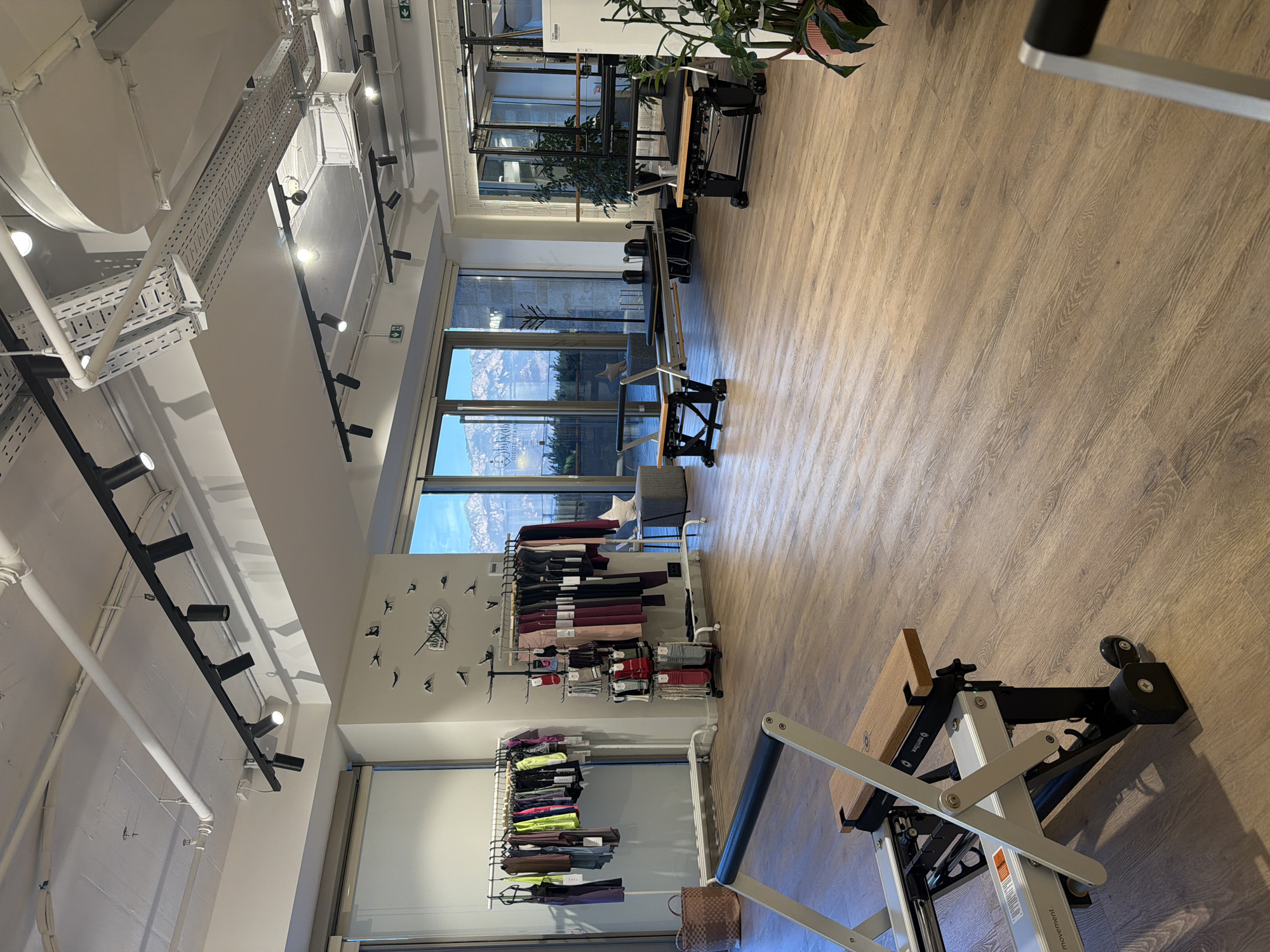 The image depicts the interior of a brightly lit Pilates studio located in the city of Boreti, Montenegro. The studio features a spacious, open-plan layout with light-colored wooden flooring. Several Pilates reformers are visible, arranged on the floor, with one in the foreground and others in the mid-ground and background. Large windows line one wall of the studio, offering a view of mountains and a clear sky, suggesting a scenic location.
In the background, racks of athletic apparel, including leggings and tops, are displayed, indicating that the studio may also sell merchandise. A decorative clock featuring silhouettes of people in yoga poses adorns one wall, along with a logo that reads "Ballet Studio". Various plants are strategically placed around the room, adding a touch of greenery. The ceiling is equipped with a modern track lighting system and industrial-style ventilation ducts. The overall atmosphere of the studio is clean, organized, and inviting, designed for physical activity and wellness. There are no people visible in the image.