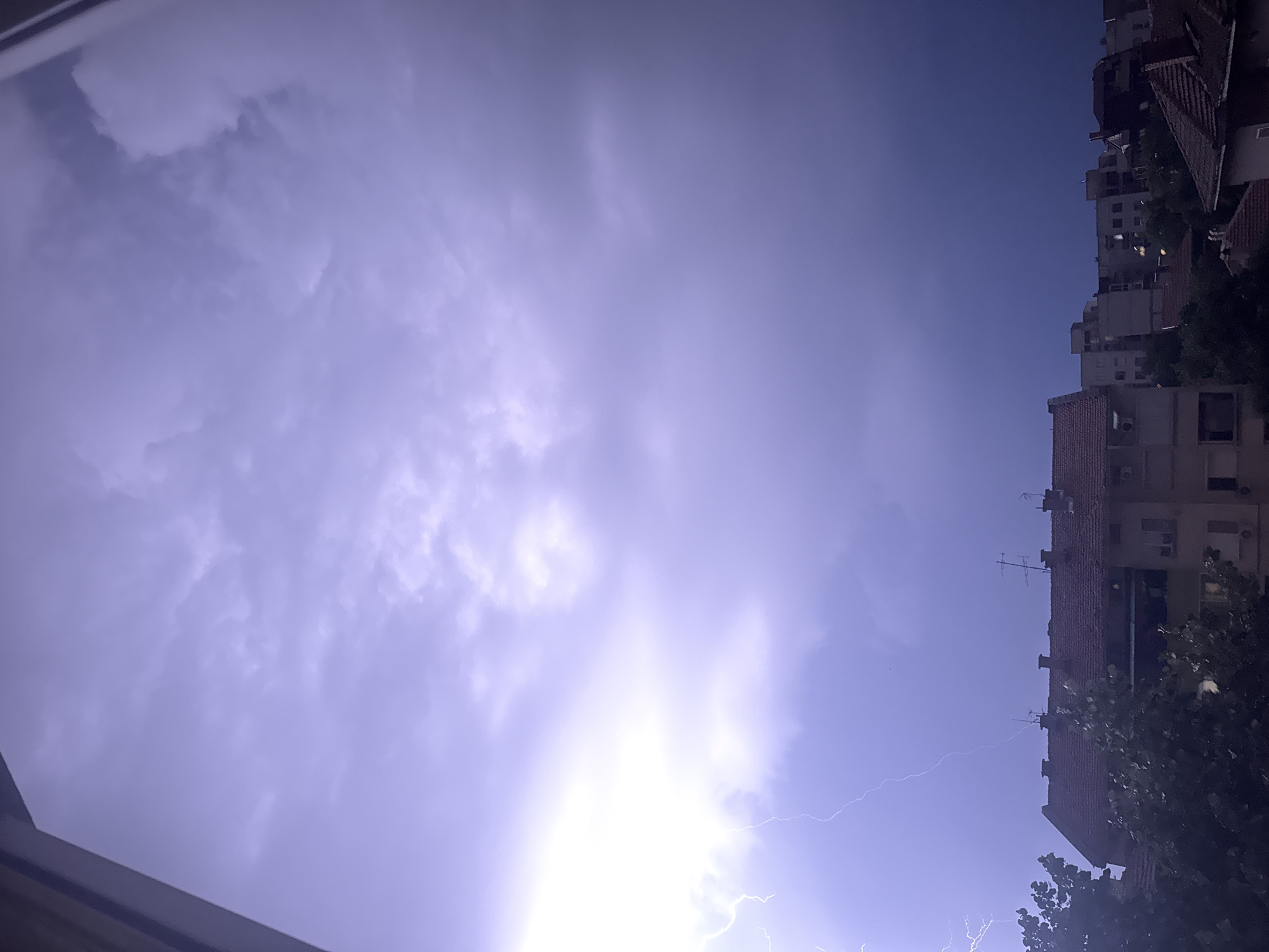The image shows a dramatic sky with a lightning strike over the city of Plovdiv, Bulgaria. The sky is filled with dark, heavy clouds, tinged with purple and blue hues, indicating an impending storm. A bright flash of lightning illuminates the clouds on the left side of the frame, with faint branching tendrils extending downwards. In the foreground, silhouetted against the stormy sky, are the rooftops of buildings. Some of these buildings appear to be apartment blocks, with multiple windows and balconies visible. Chimneys and television antennas dot the rooftops. Trees with dark foliage are also present in the lower part of the image, adding to the sense of an approaching storm. The overall impression is one of natural power and urban resilience.