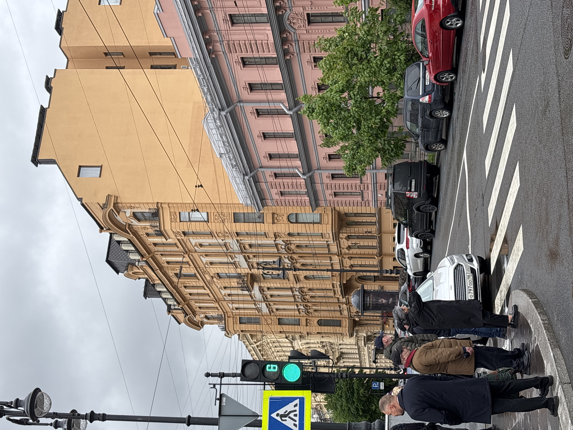 The image depicts a street scene in Saint Petersburg, Russia. Buildings with ornate facades line the street, featuring a mix of colors from beige to pink. A row of parked cars, including red, black, and white vehicles, are visible along the edge of the road. A pedestrian crosswalk is in the foreground with pedestrians waiting to cross the street. A traffic light is green, and a crosswalk sign is visible. Streetlights line the edge of the street.