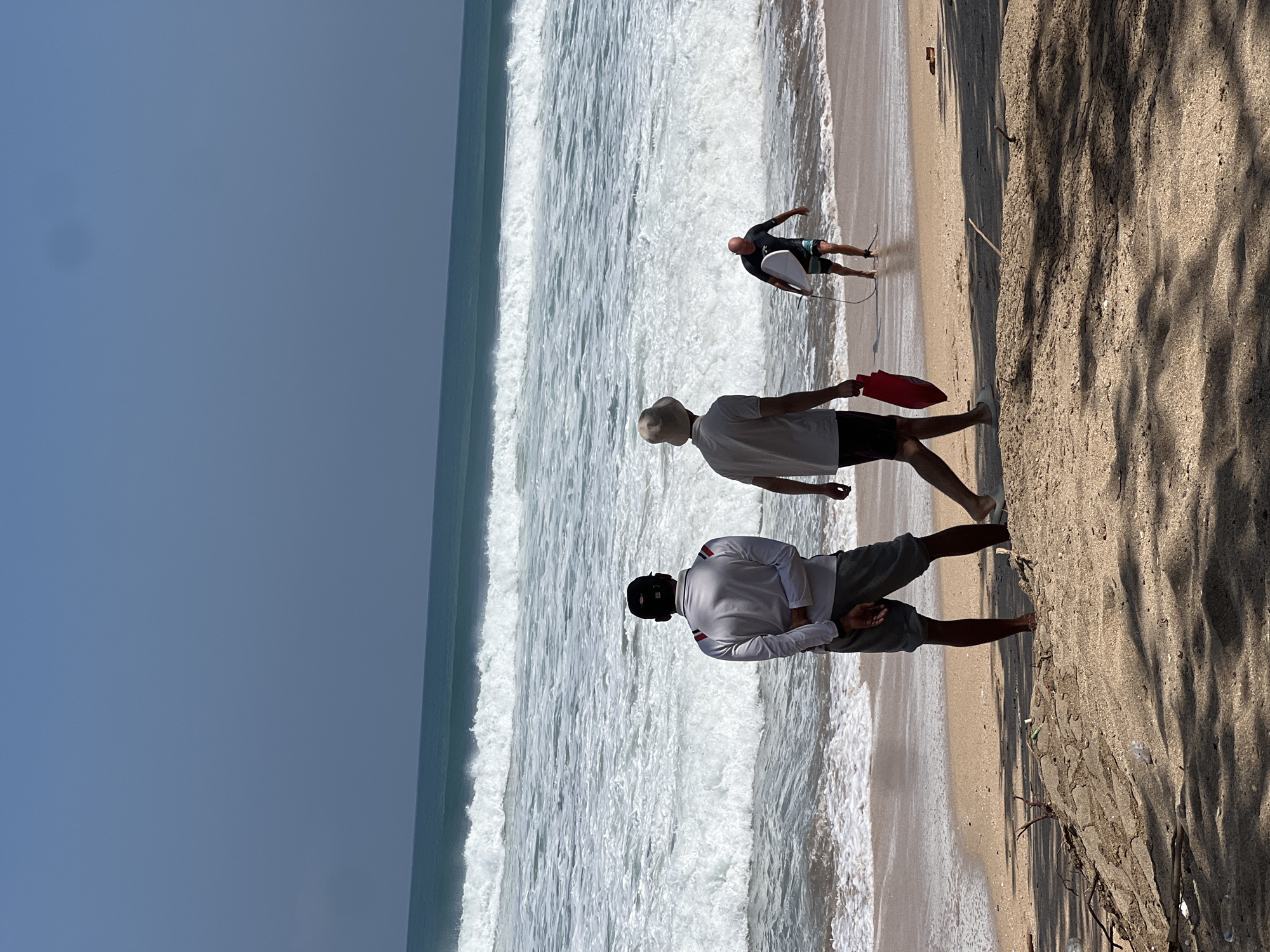 A sunny day on a beach in Kuta, Indonesia, features three individuals near the shoreline with crashing waves in the background. In the foreground, two men stand with their backs to the camera, facing the ocean. The man on the left wears a white long-sleeved shirt and dark shorts, with his hands clasped behind his back. The man to his right wears a light-colored bucket hat and a white short-sleeved shirt, carrying a red bag in his right hand. Further down the beach, a third man, partially obscured by distance and the other two, is entering the water with a surfboard under his left arm. He is wearing dark shorts and a dark t-shirt. The sand is a light brown, with scattered debris and tree shadows cast across it. The ocean exhibits significant wave activity with white foam cresting. The sky above is a clear, light blue.