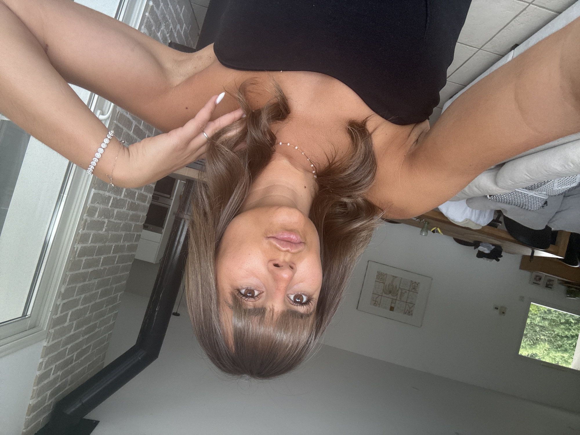 A woman wearing a black top takes a selfie in an interior space. She has brown hair with bangs and is wearing jewelry on her wrist and neck. In the background, there is a window, a brick wall, a wood-burning stove, a piece of art on the wall, and a window with greenery visible outside. The location is Veauchette, France.