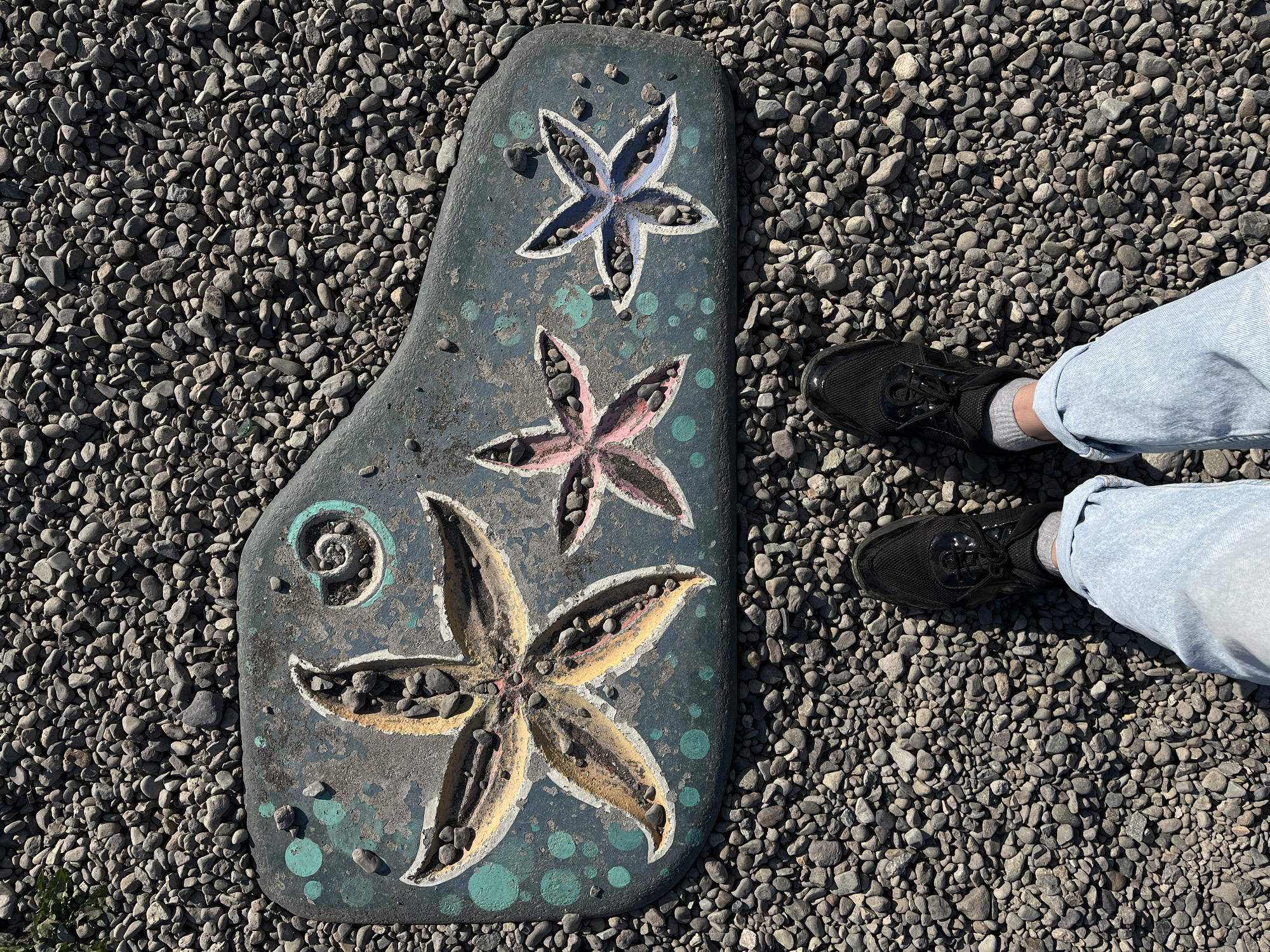 An overhead perspective captured in Petropavlovsk-Kamchatsky, Russia, depicts an irregular, dark teal-colored decorative slab positioned on a field of small, grey, rounded gravel. The slab is adorned with three painted starfish motifs, each recessed and filled with pebbles identical to the surrounding ground. The largest starfish, on the left, is painted yellow and orange; the central starfish is pink; and the smallest starfish, on the right, is blue and purple. All starfish are edged with white paint. A teal spiral design and scattered teal dots are also painted on the slab. At the bottom of the frame, the lower legs and feet of an individual are visible. The individual wears light-wash blue denim jeans, cuffed above the ankles, grey socks, and black athletic shoes with black laces. The scene is illuminated by direct sunlight, evidenced by shadows cast on the gravel and slab.