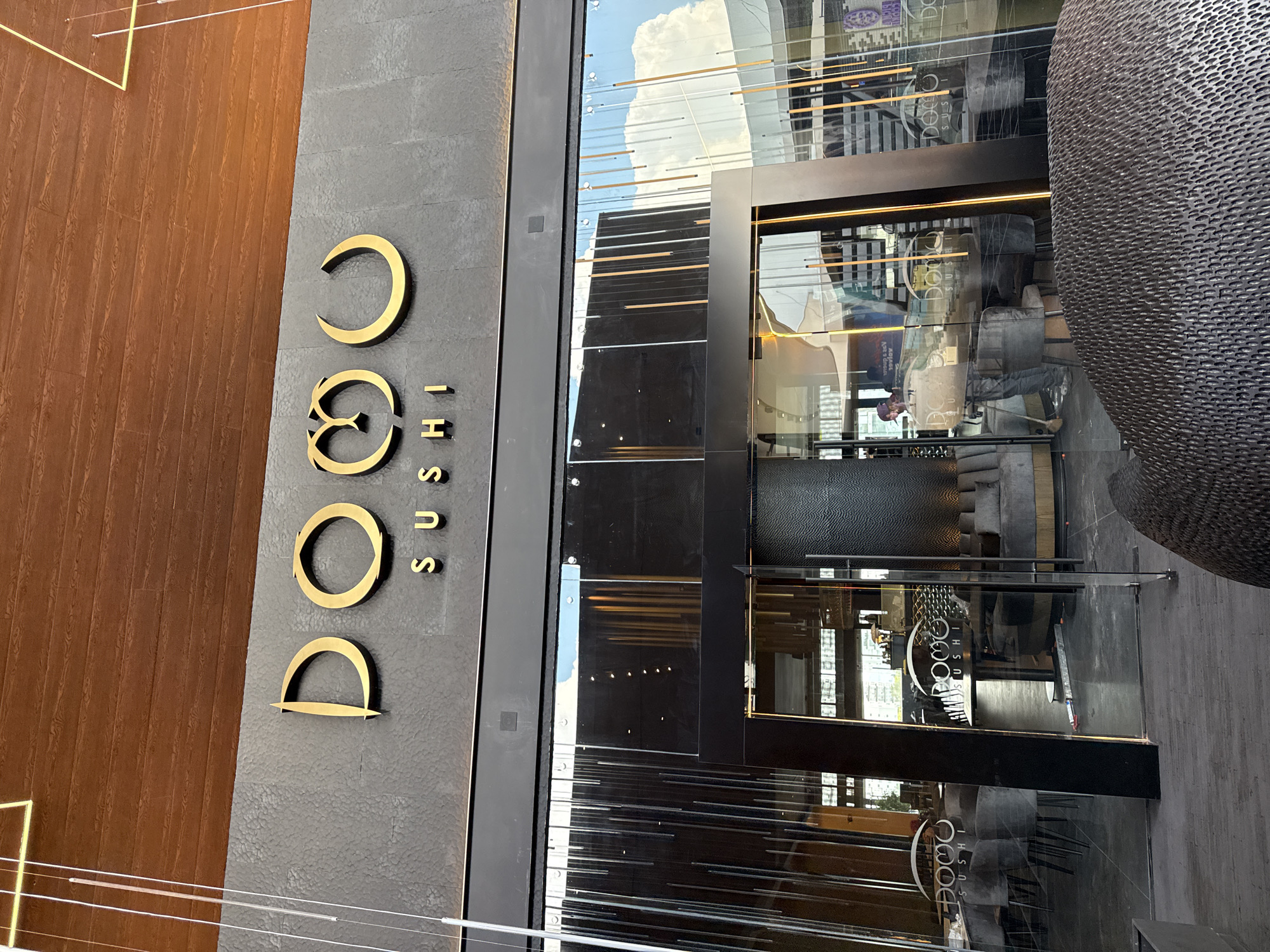 The image shows the entrance of a sushi restaurant called "DOMO Sushi". The name is vertically displayed on a dark gray wall in gold lettering. To the left of the wall is a brown wooden panel. The entrance consists of a glass door and windows reflecting the sky and some interior elements. Inside the restaurant, tables and chairs are visible. A large, rounded, textured structure is partially visible on the right. The photo appears to be taken outdoors, during daytime, in San Bernardino Tlaxcalancingo, Mexico. No people are prominently visible, although reflections in the glass suggest the presence of individuals inside.