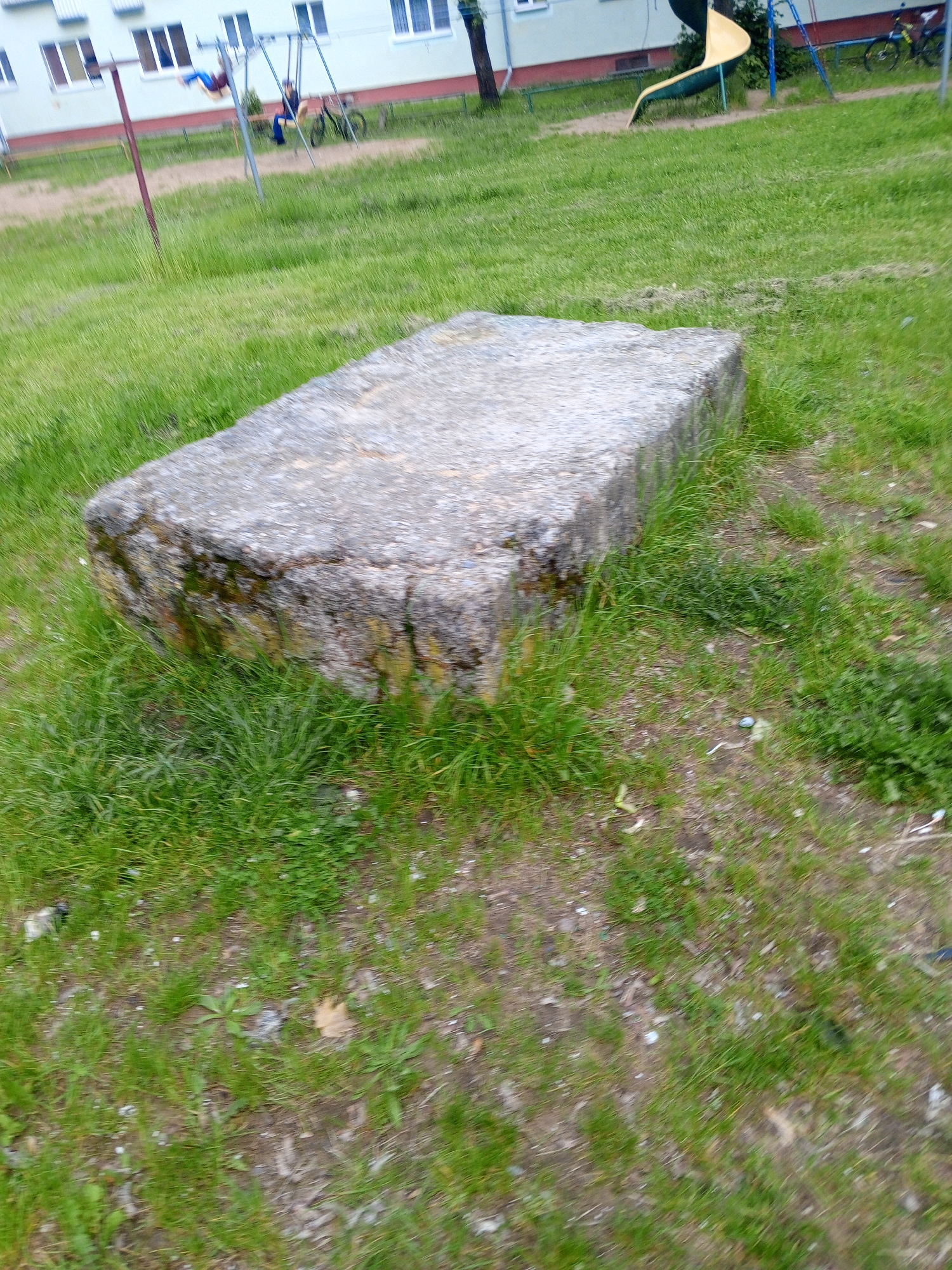The image depicts an outdoor scene, likely a public common area or playground adjacent to a residential building, located in Unknown, Unknown.
In the foreground, a large, rectangular, weathered concrete slab occupies the lower central and right portions of the frame, resting on a patchy ground of green grass and bare soil. The sides of the slab exhibit dark staining or moss growth. The ground immediately around the slab and extending into the lower frame is a mix of green grass, sparse dirt, and scattered small debris.
In the midground, towards the left, a light-colored multi-story building with several windows is visible. The windows have white frames. In front of the building, a metal swing set is present. One child, largely blurred by motion, is actively swinging high, wearing blue pants and a dark top. Another child is seated on a swing to the right of the first, wearing blue pants and a dark top, appearing more static. A bicycle is parked near this child. A reddish-brown pole stands vertically on the far left. Towards the right midground, a yellow, curved playground slide is visible, partially obscured by a dark tree trunk. The entire scene is under diffused or overcast lighting conditions, with no strong shadows evident. Motion blur is pronounced on the swinging child and the surrounding grassy areas.