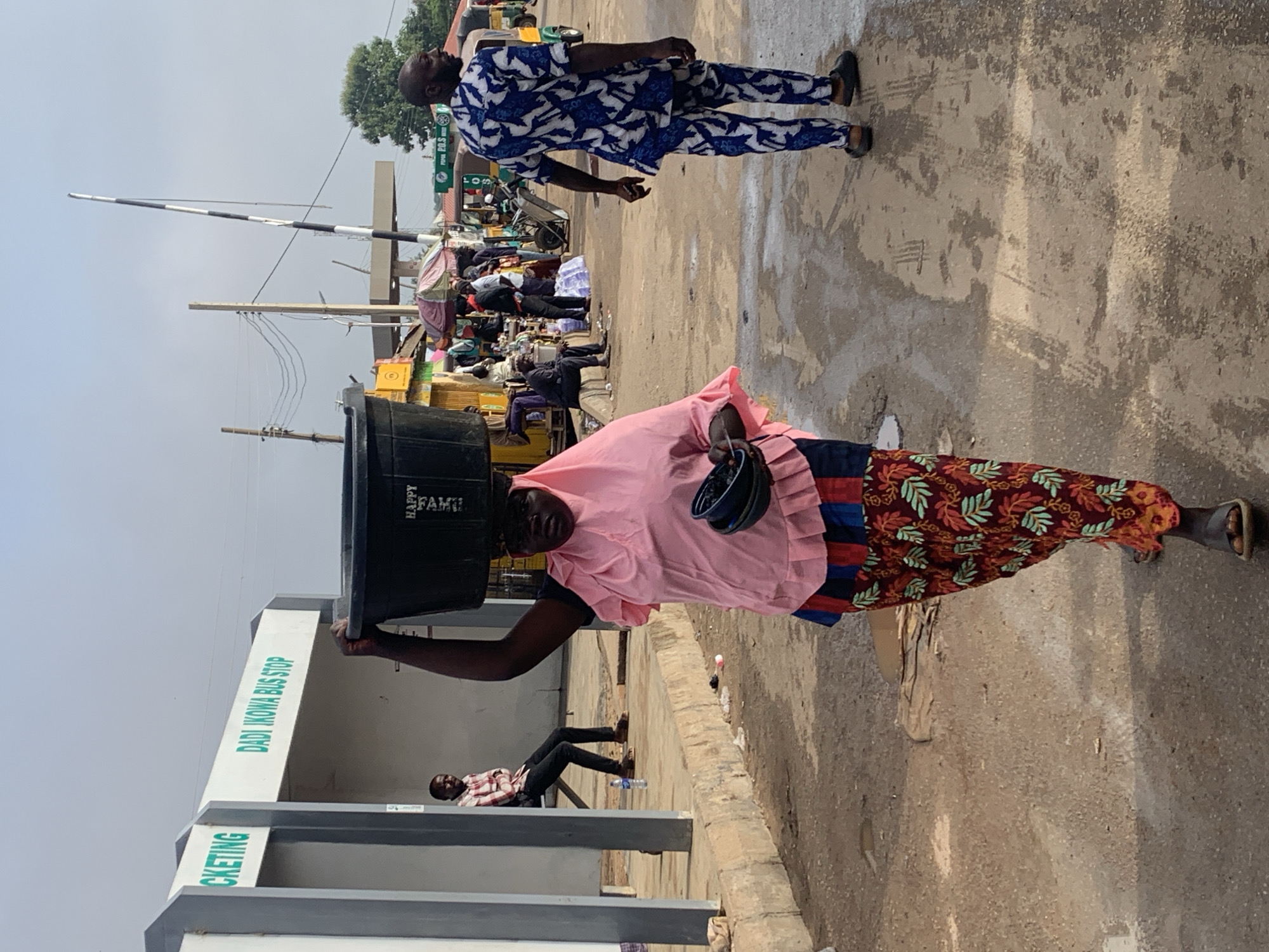 In Gura Top, Nigeria, a woman wearing a pink hijab and a colorful patterned skirt walks carrying a large black bucket on her head and two smaller dark bowls in her right hand. She is walking on a dusty, wet road in front of a bus stop labeled "DADI IKOWA BUS STOP." To her left, a man in a plaid shirt and dark pants is seated. To her right, a man in a blue and white patterned traditional outfit walks away from the camera. In the background, there are other people, market stalls, a barrier gate, and utility poles. The sky is overcast.