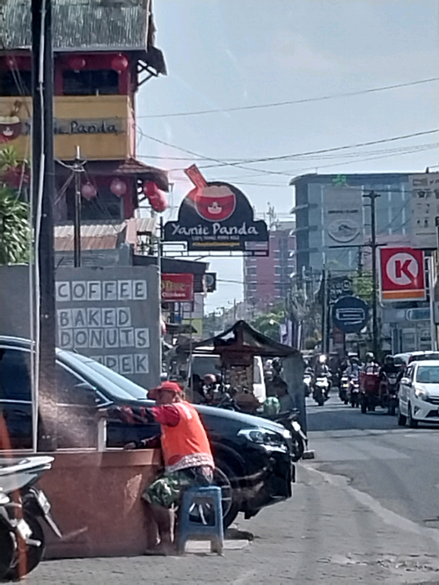 A street scene in Depok, Indonesia, shows a person in an orange vest and patterned shorts sitting on a blue stool next to a dark-colored vehicle. The vehicle appears to be a car, with its front passenger side visible. Behind the vehicle is a concrete structure with text on it that reads "COFFEE BAKED DONUTS". A street vendor's cart with a brown counter and a blue opening is also present. In the background, there are several motorcycles and cars on a road, indicating traffic. Buildings with signs are visible on the right side of the street, including a sign for "Yamie Panda". The sky is clear and blue.