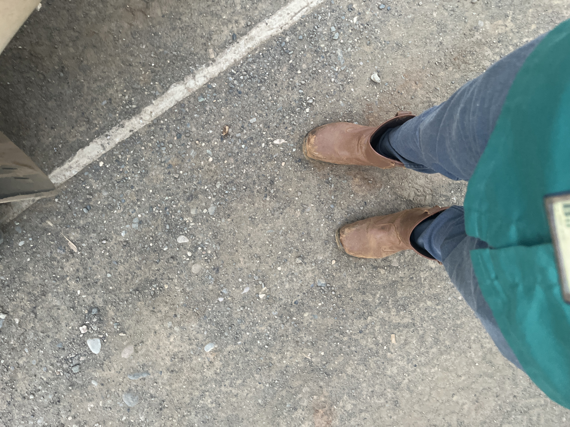 The image shows a person's legs and feet wearing brown boots and blue jeans, standing on a gravel surface. A white line, likely a parking lot marking, is visible on the right side of the image. A portion of a dark green garment is visible in the lower part of the frame. The overall scene appears to be outdoors in an urban or semi-urban environment, such as a parking lot or a street. The lighting suggests it is daytime. No specific details indicate the location in the city of Unknown, Indonesia.