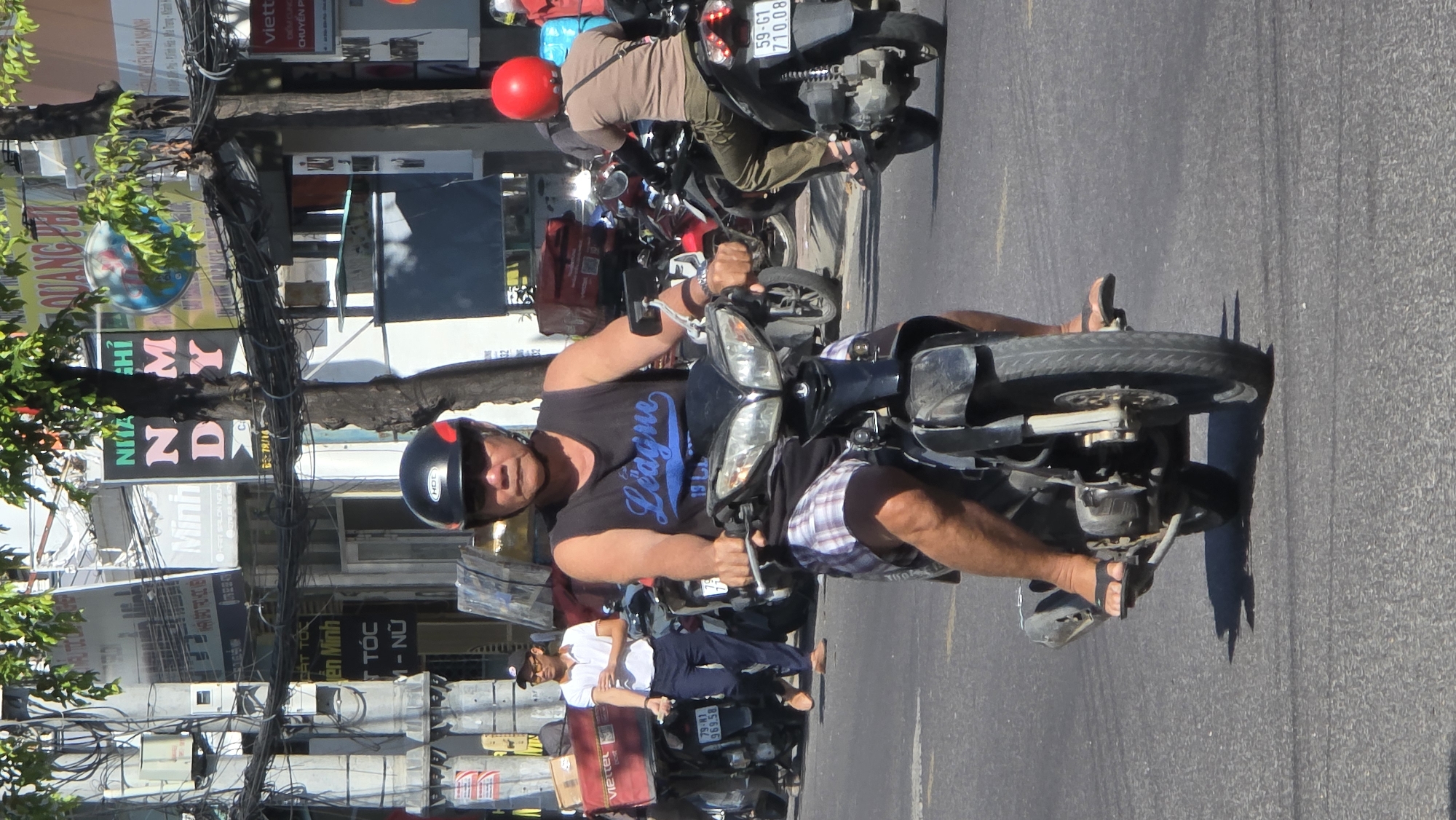 The image shows a man driving a motorcycle on a street in what appears to be Bắc Nha Trang, Vietnam. The man is wearing a black helmet, a black t-shirt with "League" written on it, and checkered shorts. He is also wearing sandals. Behind him, another person with a red helmet is riding a motorcycle. Street signs written in Vietnamese are visible, along with storefronts and overhead electrical wires. The street is paved and sunny. Several other parked motorcycles are also visible.