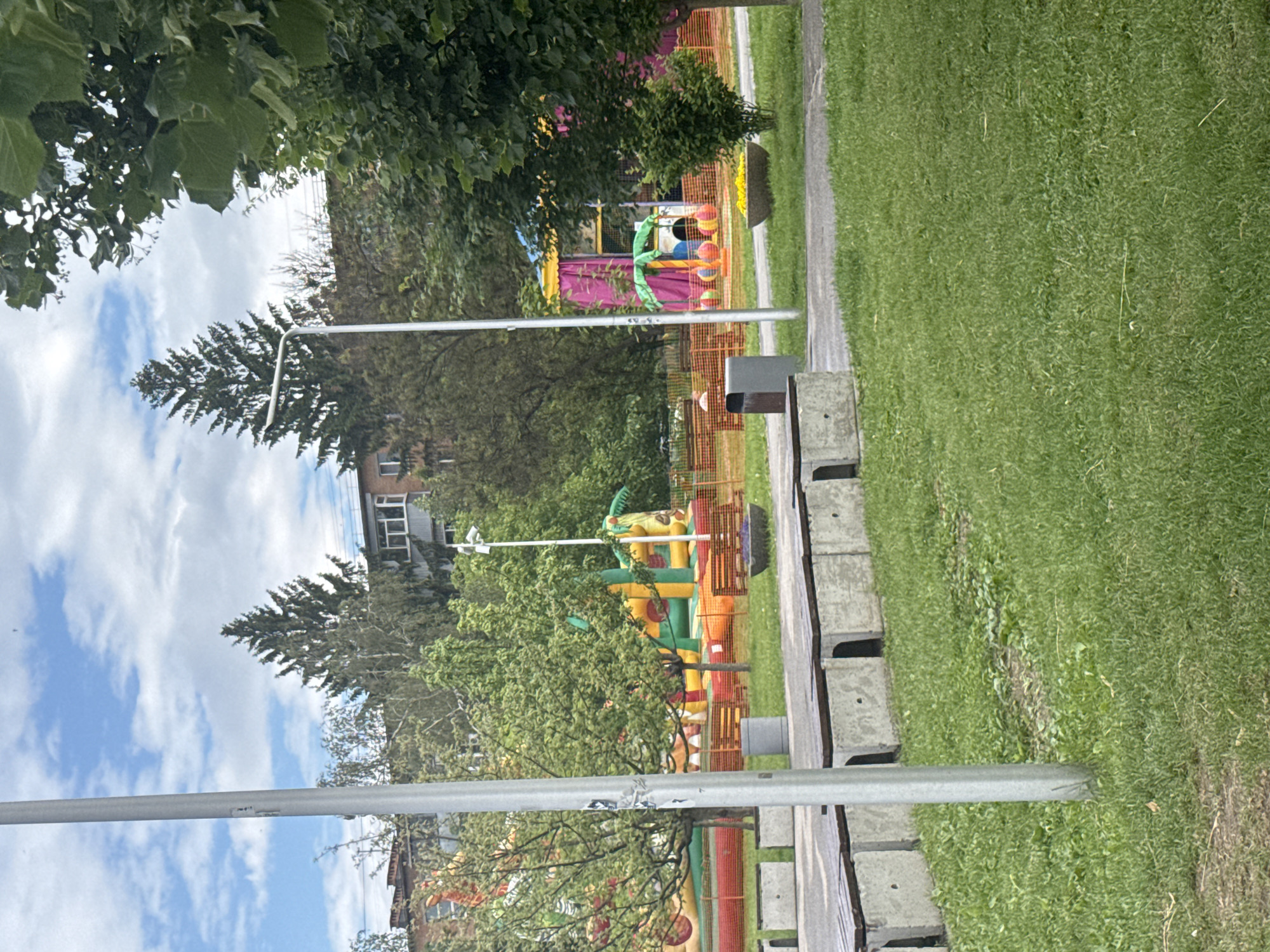The image captures an outdoor scene featuring a colorful inflatable playground situated within a park-like setting. In the foreground, a grassy area with a dirt or gravel path runs along the right side, bordered by concrete benches. Several tall, slender metal poles, likely streetlights or utility poles, punctuate the scene vertically.
The central focus is a vibrant inflatable playground, enclosed by an orange safety net or fence. The equipment includes multiple large bouncy structures in various bright colors such as yellow, green, pink, purple, and red. One inflatable appears to be shaped like a green dinosaur or dragon, while another features a palm tree design. Smaller colorful inflatable balls or obstacles are also visible within the play area.
Behind the playground, dense green trees and foliage are present, partially obscuring multi-story buildings in the distance, suggesting a suburban or urban park environment. The sky above is partly cloudy with patches of blue. The overall lighting indicates it is daytime. No people are clearly visible engaging with the playground equipment, or in the wider scene. There is no discernible text visible in the image.