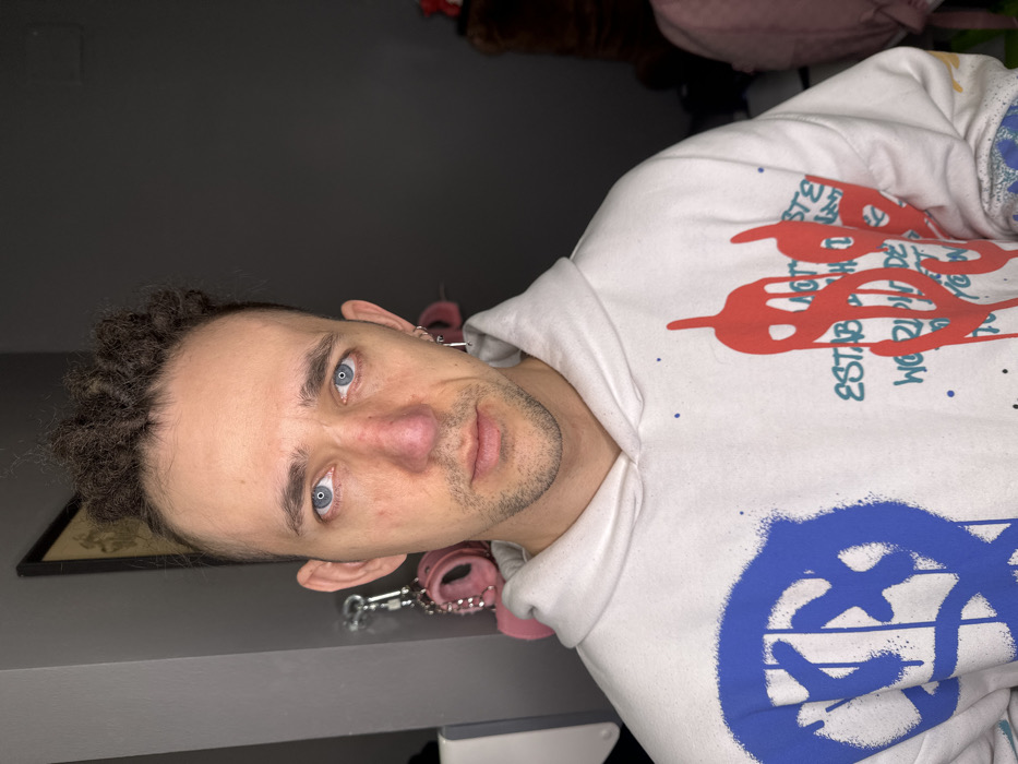 The image presents a male individual, appearing between 20 and 40 years of age, captured from the chest up in a frontal view, gazing directly at the camera. He has blue eyes and a prominent nose with redness visible on the bridge and tip. His hair features dark, curly dreadlocks on the crown with shaved sides, and a receding hairline. His left ear displays multiple silver-toned hoop and bar earrings. He is wearing a white hooded sweatshirt adorned with graffiti-style graphics: a large blue spray-painted circle with crossed lines on the left chest, and a large red dripping dollar sign symbol on the right chest, complemented by smaller text and splatters in various colors. A blue and white patterned cuff is visible on his right wrist.
The indoor background consists of a dark gray wall. To the subject's left, a white rectangular object with a dark vertical line is visible. Hanging from a fixture, beneath a dark-framed picture, is a pair of pink, fur-covered handcuffs with silver-colored chains. To the subject's right, indistinct shadows and partially discernible faint pink and brown soft objects are present.
The specific location within the city of Saratov, Russia, cannot be determined from the visual information provided.