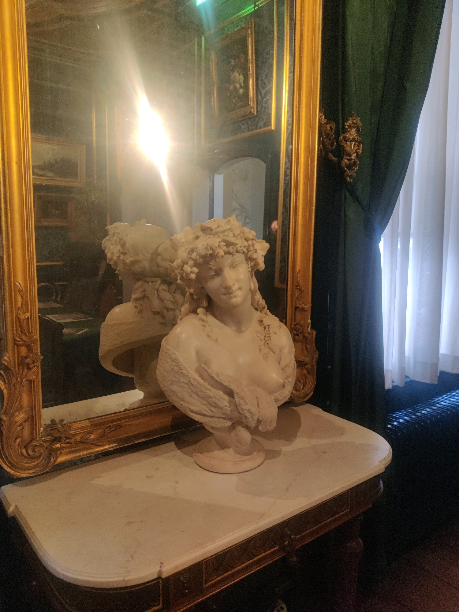 The image shows a marble bust of a woman with a floral headdress, placed on a marble-topped table. The bust is reflected in a large, ornate gilded mirror. Behind the mirror, dark green curtains are partially drawn, revealing sheer white curtains. The room appears to be dimly lit, with a strong light source reflecting off the mirror, obscuring details in the background reflections. The table has a decorative wooden base with intricate carvings. The scene is devoid of any people or any indication of activity. The location is not discernible from the image itself, and thus cannot be identified as Amsterdam, Netherlands.