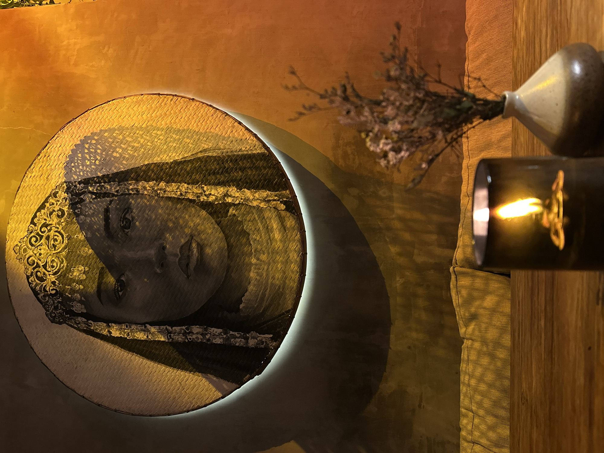 An eye-level, indoor shot taken in Pererenan, Indonesia, features a room corner with warm lighting. On the wall, a circular woven artwork displaying a woman's face with intricate head adornments is mounted, featuring a subtle illuminated ring outlining it. Next to the artwork, to the right, is a small vase holding dried flowers, placed atop what seems to be the corner of a sofa. A lit candle in a dark container sits to the right of the vase. The background comprises a textured wall with an orange hue gradient.