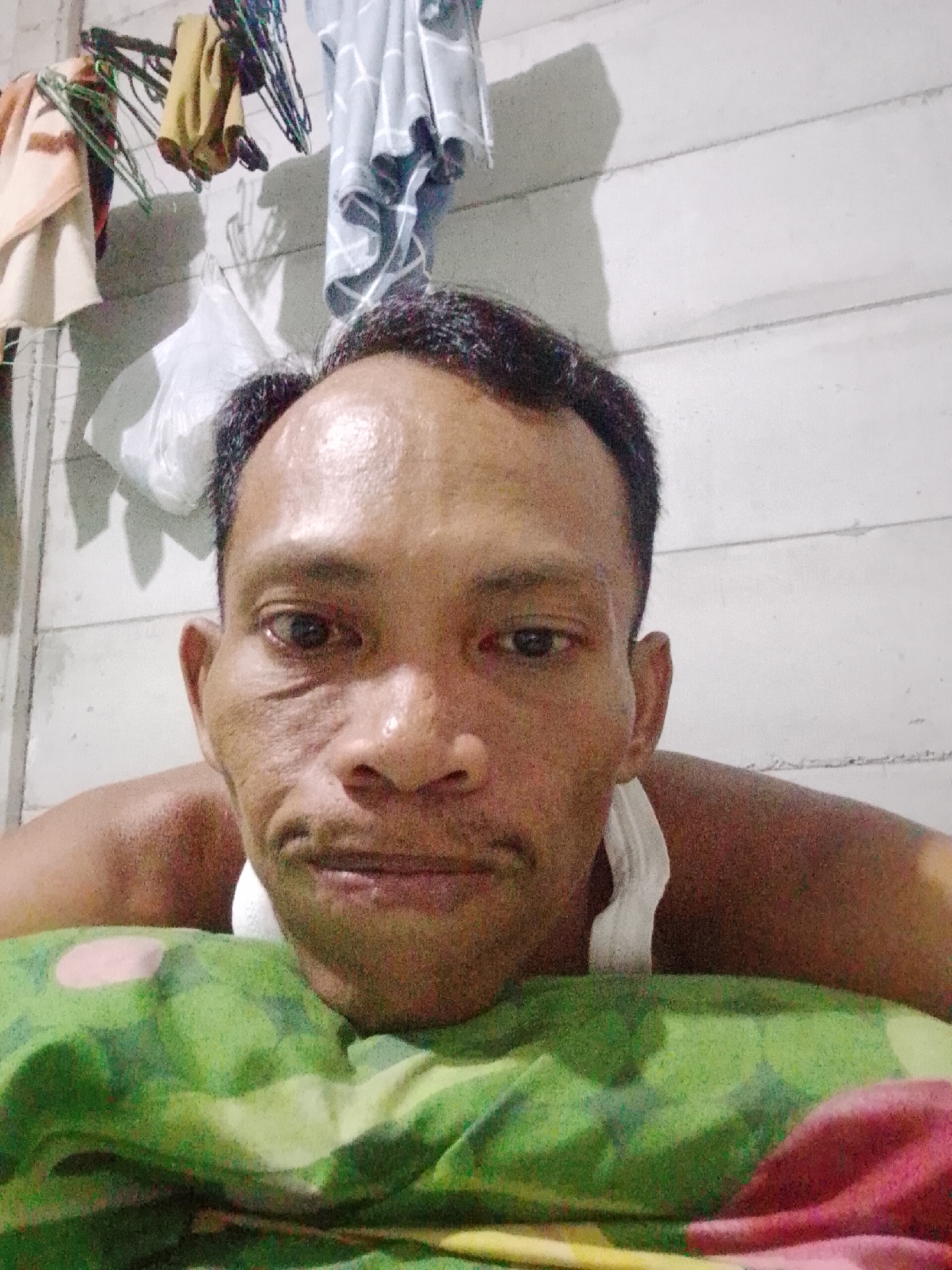 The image depicts a man lying down with his face close to the camera, resting on a green and red patterned pillow. He appears to be wearing a white tank top. In the background is a white wall with clothing hanging on hangers. The image was presumably taken indoors in Bukit Ketok, Indonesia.