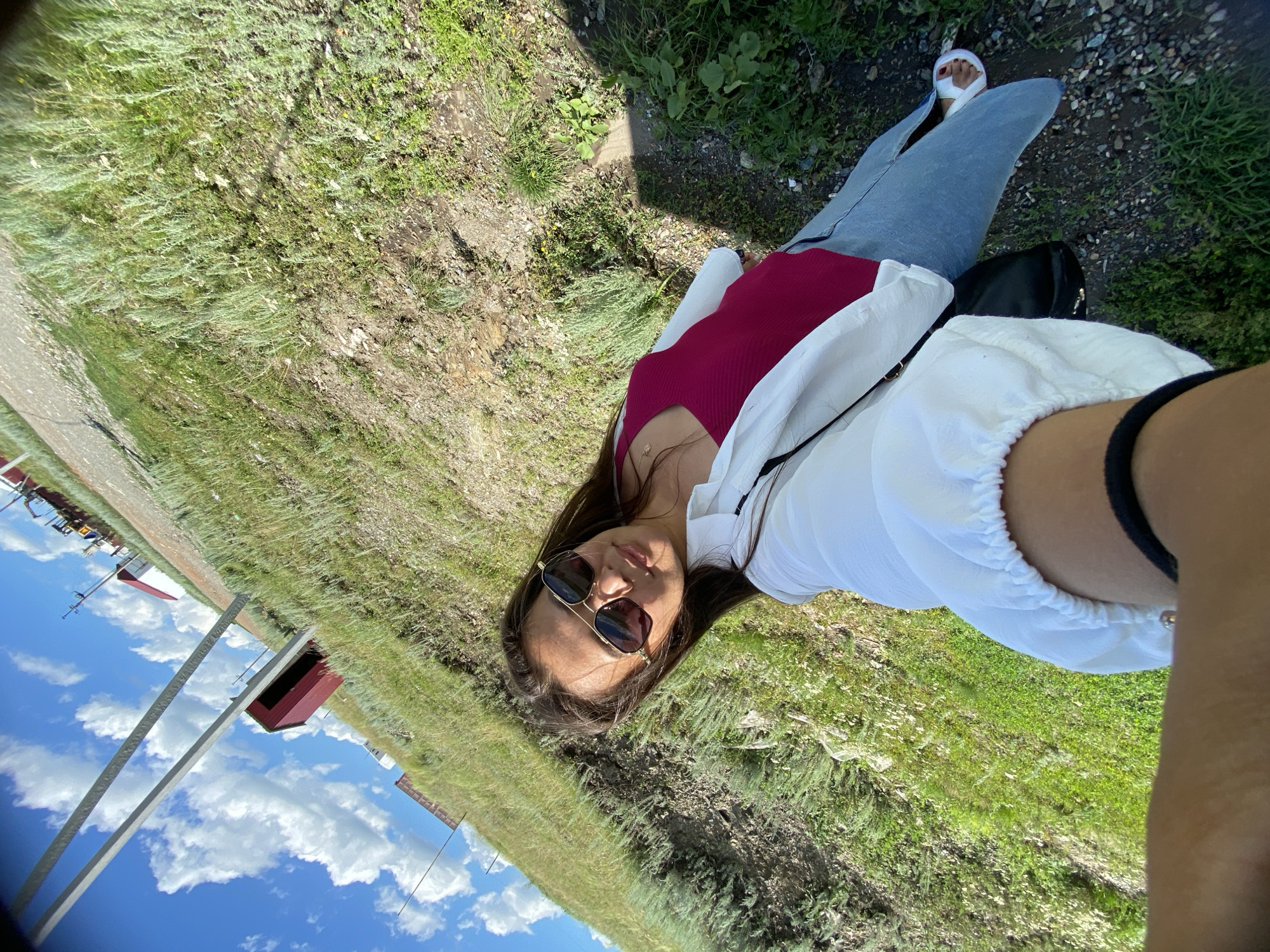 A woman, wearing sunglasses, a fuchsia tank top, a white button-up shirt, and light blue jeans, is taking a selfie outdoors on a sunny day. She is standing on a grassy, hilly terrain with scattered rocks and dry vegetation. In the background, there is a gravel road, a red building, utility poles, and distant industrial equipment. The sky is blue with scattered white clouds. The scene appears to be in a rural or semi-industrial area.