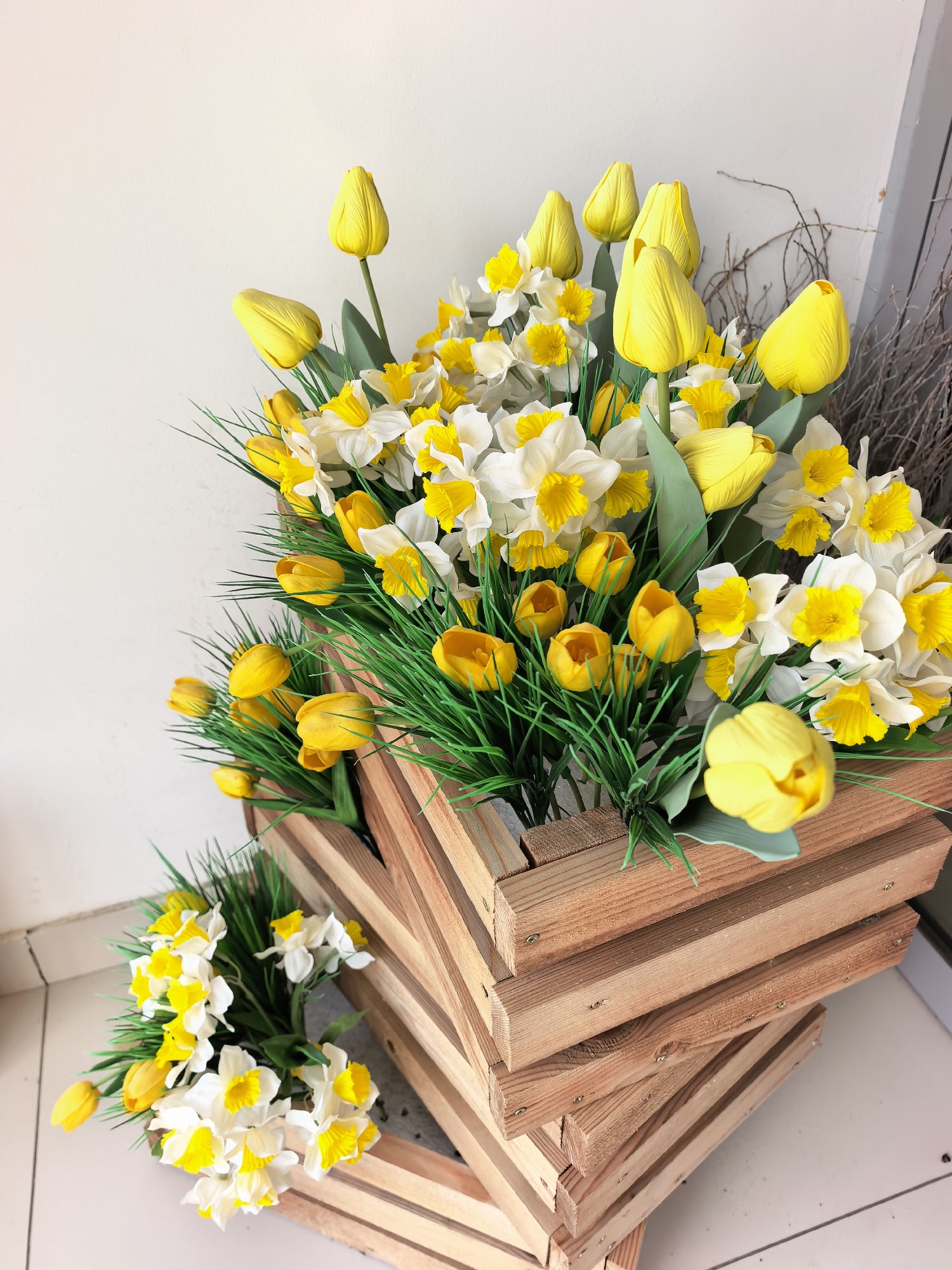 The image depicts a rustic wooden planter filled with an abundant arrangement of artificial yellow tulips and white and yellow daffodils, interspersed with artificial green grass. The planter itself is constructed from light-colored wood planks and appears to be stacked or designed in a spiral fashion, giving it a dynamic visual element. The flowers are densely packed and vibrant, suggesting a celebratory or decorative display. The background consists of a plain, light-colored wall and a floor that appears to be tiled, with a hint of a doorway or opening on the right side, where some dry branches are visible. The lighting is even and diffused, indicating an indoor or shaded outdoor setting. The scene is static, with no people or animals present. The photograph is taken from a slightly elevated, angled perspective, focusing on the floral arrangement and the unique planter. The location is not specified in the image itself, but the prompt requests it to be within Sterlitamak, Russia. Therefore, this image is presented as if it were taken in Sterlitamak, Russia.