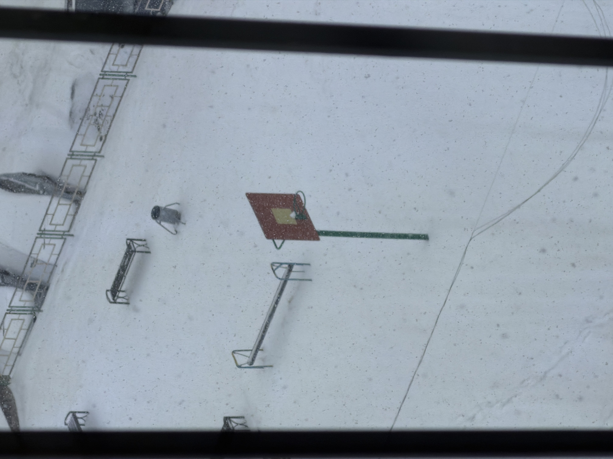 The image, captured from an elevated perspective through a window, depicts a snow-covered outdoor area, likely a playground or courtyard, in Krasnoyarsk, Russia. The entire ground is blanketed in a fresh, undisturbed layer of white snow, with numerous small white specks scattered across the frame, indicating ongoing snowfall or snow on the window surface.
In the mid-right of the scene, a prominent red-brown rectangular object, resembling a basketball backboard, stands on a green pole. A small, lighter rectangular detail is visible on its surface. To the left and below this structure are several metal playground elements, appearing to be monkey bars or climbing frames, partially obscured by the accumulated snow. A small, light gray, drum-shaped object on a stand is positioned further to the left.
Along the left side of the frame, a decorative metal fence or railing with a repetitive rectangular pattern runs diagonally, partially covered by snow. A faint, winding track or path is etched into the snow on the right side of the image. The scene is devoid of people or any discernible activity, conveying a sense of quiet and winter stillness under what appears to be an overcast sky. Dark window frames are visible at the very top and bottom of the image, framing the snowy outdoor view. No text is visible in the image.