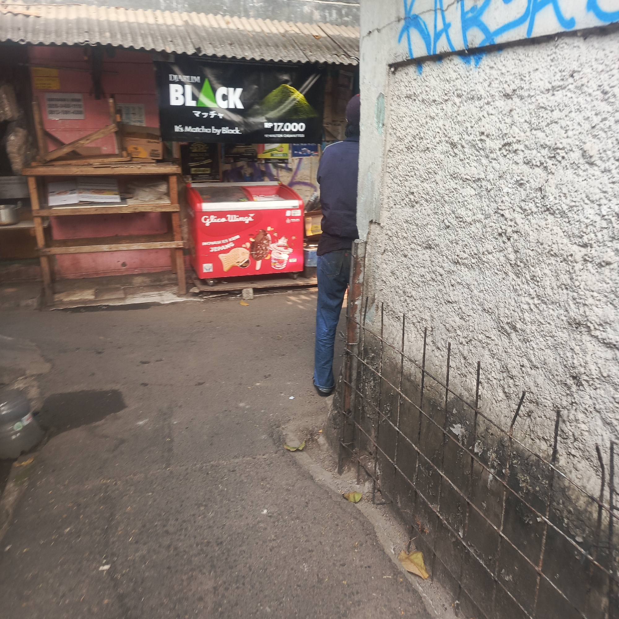 The image depicts an outdoor scene, likely on a street or alleyway in South Jakarta, Indonesia. In the background, there is a small stall or shop with a corrugated metal roof. A black banner with green text "BLACK MATCH" and "Rp 17.000" is prominently displayed. To the left of the banner, there is a wooden shelf displaying some items. In front of the stall, a red freezer chest with "Glico Wings" branding is visible, containing ice cream products. On the right side of the image, a portion of a textured concrete wall with blue graffiti is seen. A person wearing blue jeans and a dark shirt is partially visible, standing near the wall. The ground is paved with asphalt and shows signs of wear and tear, with some debris and fallen leaves. The overall impression is of a casual, everyday scene in an urban environment.