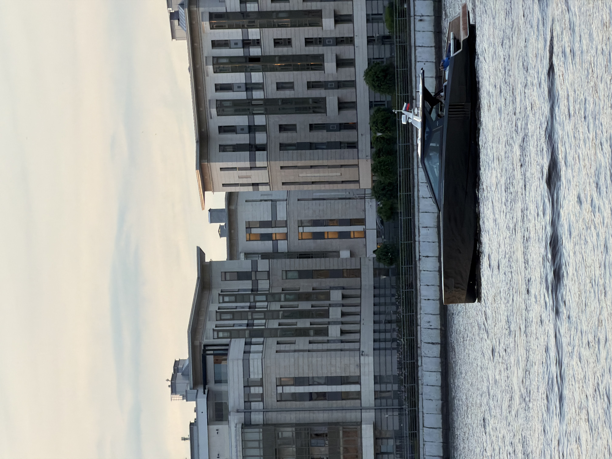 A modern, dark-hulled motor yacht with a white radar arch and a Russian flag sails on a wide, choppy body of water, likely the Neva River in Saint Petersburg, Russia. The yacht features a sleek, enclosed cabin with tinted windows and a wooden swim platform at the stern. A person in a blue shirt is visible reclining on the swim platform. In the background, a row of contemporary, multi-story apartment buildings lines the riverbank. These buildings have a light-colored stone facade with repeating patterns of windows and balconies, and some windows are illuminated, suggesting it is dusk or evening. A stone embankment with a metal fence separates the water from the buildings. The sky above is overcast with soft, muted colors.