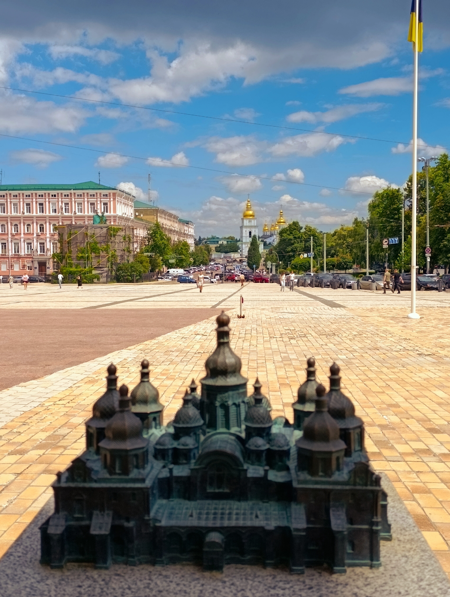 The image captures a bright, sunny day in Kyiv, Ukraine, with a clear blue sky dotted with white clouds. In the foreground, a miniature bronze model of St. Sophia's Cathedral, a prominent landmark in Kyiv, is placed on a stone base. The model features intricate details of the cathedral's domes and architecture.
The middle ground reveals a wide, paved plaza with patterned tiles. People are seen walking across the plaza. To the left, a grand, multi-story building with a green roof and ornate facade stands. Construction scaffolding with green netting is visible on a section of this building, suggesting ongoing renovations. Further down the plaza, a line of cars is parked along the right side. In the distance, the golden domes of St. Sophia's Cathedral itself are visible, rising above a cluster of trees.
On the right side of the image, a tall white flagpole stands, flying the Ukrainian flag, its blue and yellow colors a stark contrast against the sky. The environment appears to be a public square, likely Independence Square (Maidan Nezalezhnosti), given the presence of the cathedral's model and the overall urban setting. The overall atmosphere is one of a bustling city on a pleasant day.