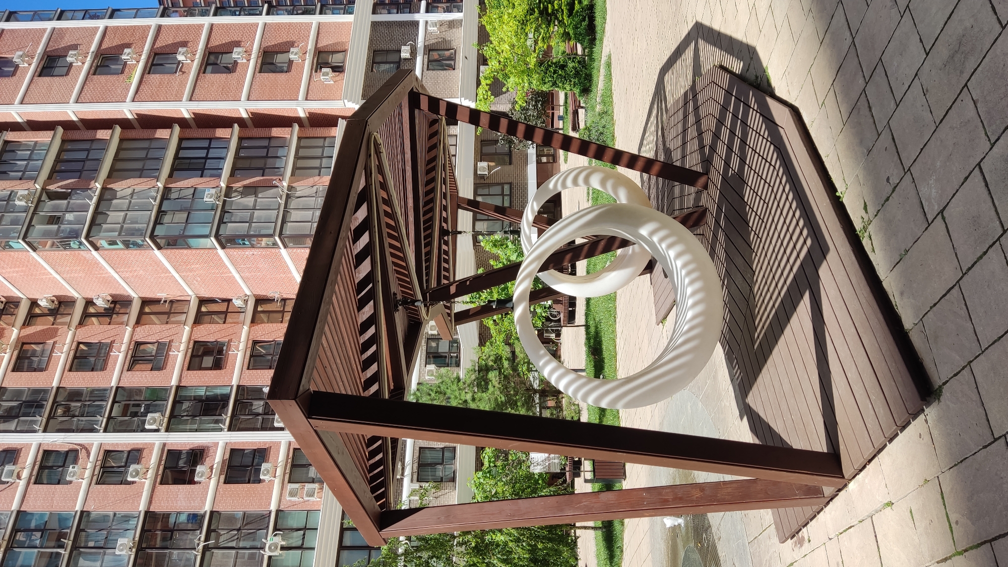 The image depicts a modern apartment building facade with brick and glass windows, situated in Krasnodar, Russia. In the foreground, a wooden pavilion with a slatted roof provides shade over a wooden deck. Two white, circular swings hang from the pavilion's structure. The surrounding area features green grass, trees, and a paved walkway. The scene is set in daylight, with shadows cast by the pavilion and swings.