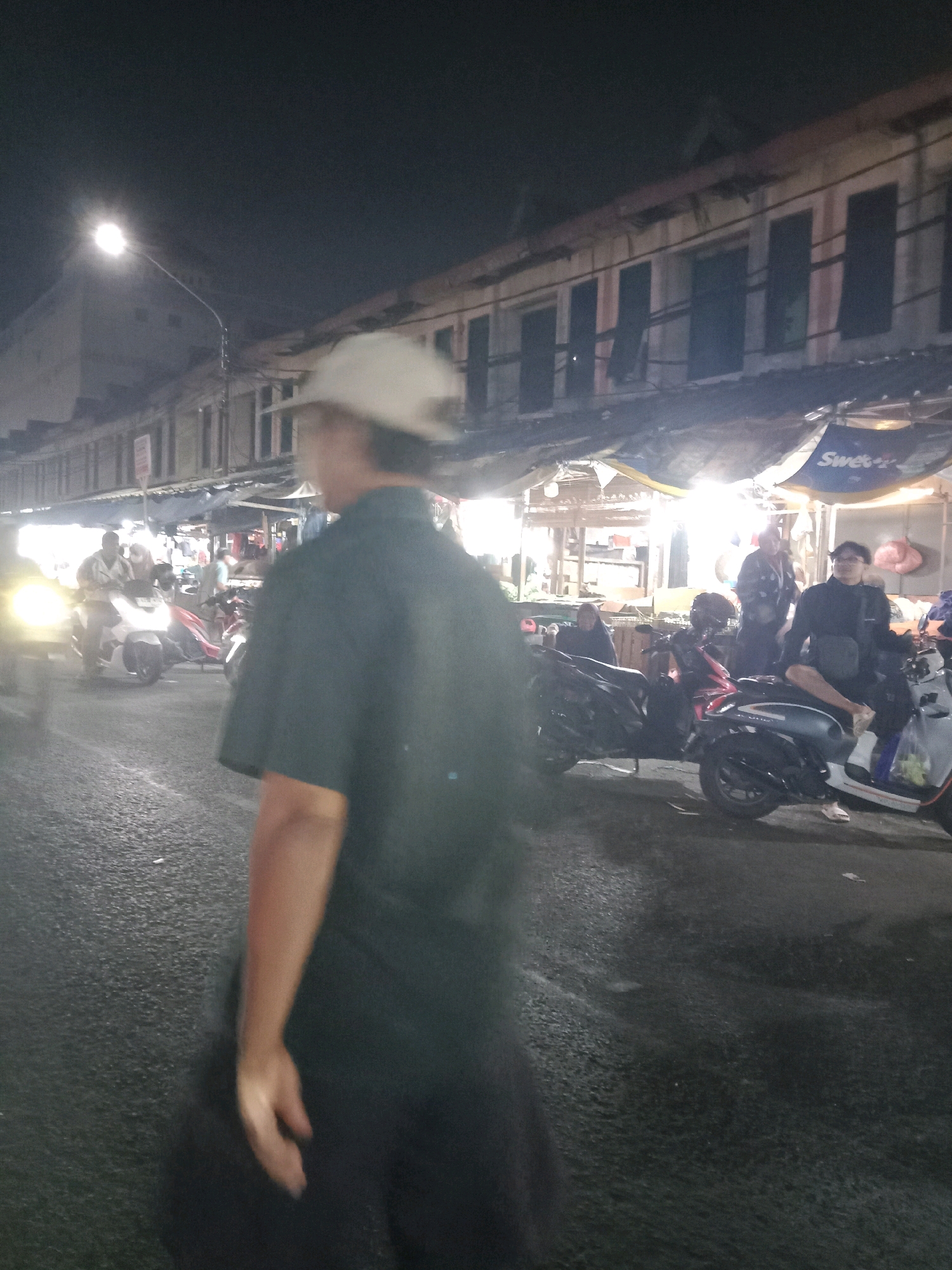 The image depicts a nighttime street scene in South Tangerang, Indonesia, likely near a market or commercial area. In the foreground, a person wearing a dark green short-sleeved shirt and a light-colored baseball cap is partially visible from the back, walking away from the camera. The street is wet, reflecting the artificial lights from nearby stalls and street lamps.
In the middle ground, several motorcycles are parked and in use. One person is seated on a light-colored scooter, wearing glasses and a dark jacket. Another motorcycle has its headlight on, with a person riding it. Further back, more motorcycles are visible, some with riders. Stalls with awnings are set up along the side of the street, illuminated by bright lights. These stalls appear to be selling various goods. The buildings in the background are multi-story structures with visible windows and signs, some of which are illuminated. The overall atmosphere is that of a bustling night market or street where commerce is taking place.