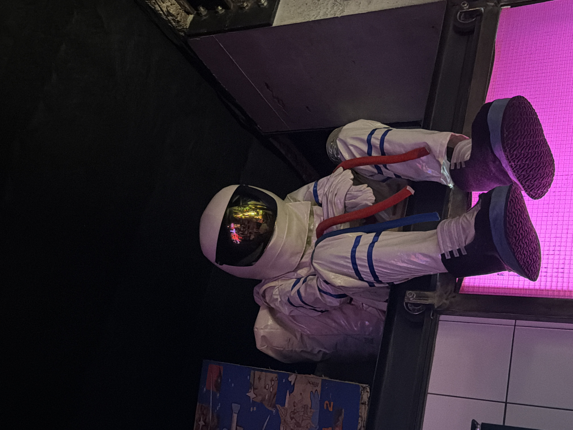 The image captures a life-sized mannequin, attired in a white astronaut suit detailed with blue stripes on the limbs and dark boots featuring blue trim and textured soles, seated on a dark, thick horizontal beam. The helmet's dark, reflective visor mirrors multi-colored light sources, predominantly warm yellow and red, indicating illuminated interior elements. Two red coiled tubes are draped across the mannequin's chest, appearing to connect to the suit. The mannequin's legs dangle downwards, positioned above a large, rectangular panel emitting a strong magenta-pink light with a visible grid pattern. To the mannequin's immediate left, a portion of a blue decorative panel with illustrations is visible. Below the beam on the left, white tiled walls are partially seen, while a dark vertical structural element is present on the right. The upper background is enveloped in darkness, suggesting an overhead space with low ambient lighting. This specific setting is identifiable as the interior of **The Space Bar** (also known as Space Cafe), located in **Sofia, Bulgaria**, where the astronaut mannequin and distinctive pink grid lighting are signature decorative elements.