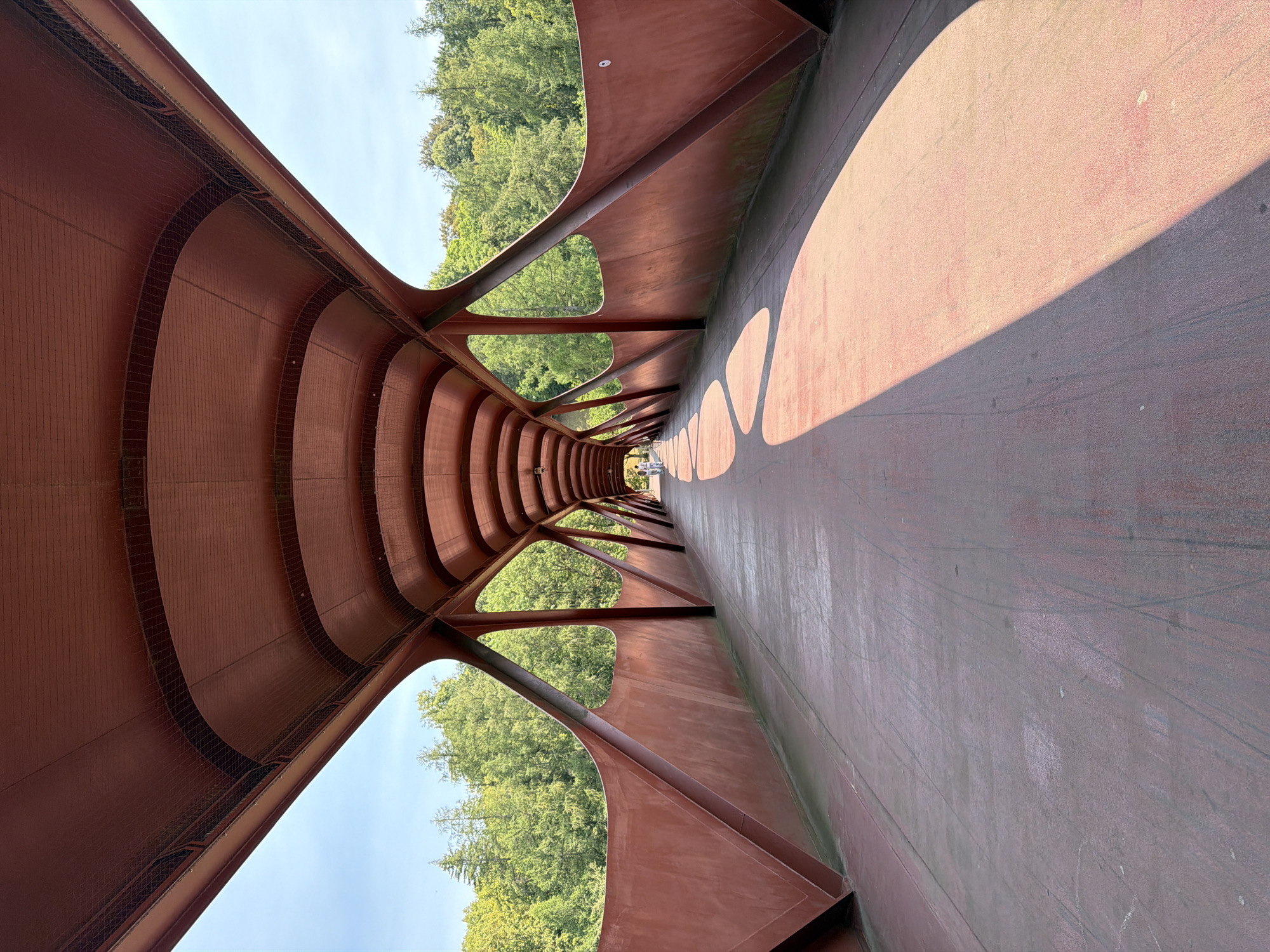 The image shows a perspective view of the interior of a covered pedestrian bridge in Esch-sur-Alzette, Luxembourg. The bridge has a distinctive, modern design with a reddish-brown exterior and a curved, vaulted roof. The roof structure is supported by a series of triangular metal beams that create open, triangular-shaped windows, offering glimpses of the lush green trees and blue sky outside. The pathway of the bridge is also the same reddish-brown color and shows some wear and tear, with visible scuff marks and faint lines. Sunlight casts dramatic shadows across the pathway, creating a stark contrast between light and dark areas. In the distance, towards the vanishing point of the bridge, two figures can be seen walking. The overall impression is one of architectural interest and a functional pathway set within a natural environment.