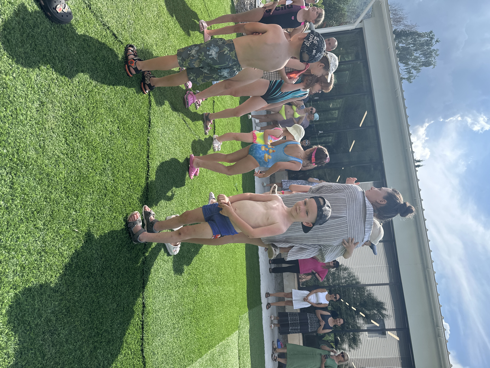 The image depicts a group of people, primarily children and adults, gathered outdoors on a sunny day. They are standing on a bright green artificial turf surface, with a building featuring large windows in the background. The scene appears to be a casual outdoor event, possibly a pool party or a gathering at a recreational facility.
Several children are wearing swimwear, suggesting they might be preparing to swim or have just finished. One young boy in the foreground is shirtless, wearing blue swim trunks and a baseball cap, and he is looking towards the camera. Other children are seen in various swimsuits, including a blue one with a duck pattern and a black and white striped one. Some adults are also present, dressed in casual clothing, with one woman wearing a striped shirt and holding a child.
The time of day is clearly indicated by the strong shadows cast on the artificial grass, suggesting it is midday or early afternoon. The sky is bright blue with fluffy white clouds, indicating fair weather. The presence of a building with large windows and what looks like a white overhang or roofline might suggest a modern facility or a resort. There is no visible text in the image. The overall mood seems relaxed and cheerful, with the individuals appearing to be enjoying themselves in the outdoor setting.