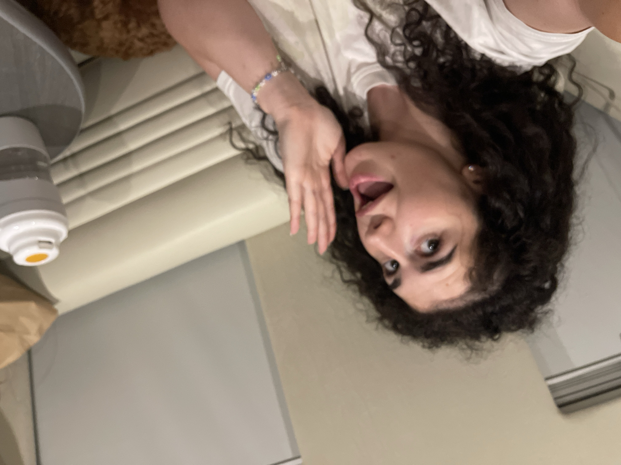 The image captures a young adult female with dark, curly hair and fair complexion, positioned centrally in an indoor setting in Koprinka, Bulgaria. She is wearing a light-colored t-shirt, a small stud earring in her left ear, and a multicolored beaded bracelet on her left wrist. Her mouth is open, and her eyes are wide, directed slightly upward and to the left of the frame. Her left hand is raised with fingers slightly splayed, positioned near her face. The background consists of light beige walls and structured seating or paneling featuring horizontal ridges. In the mid-right, a white container with an orange cap rests on a round, grey surface. A brown paper bag is partially visible behind the container. A reddish-brown, textured object is visible in the lower right quadrant.