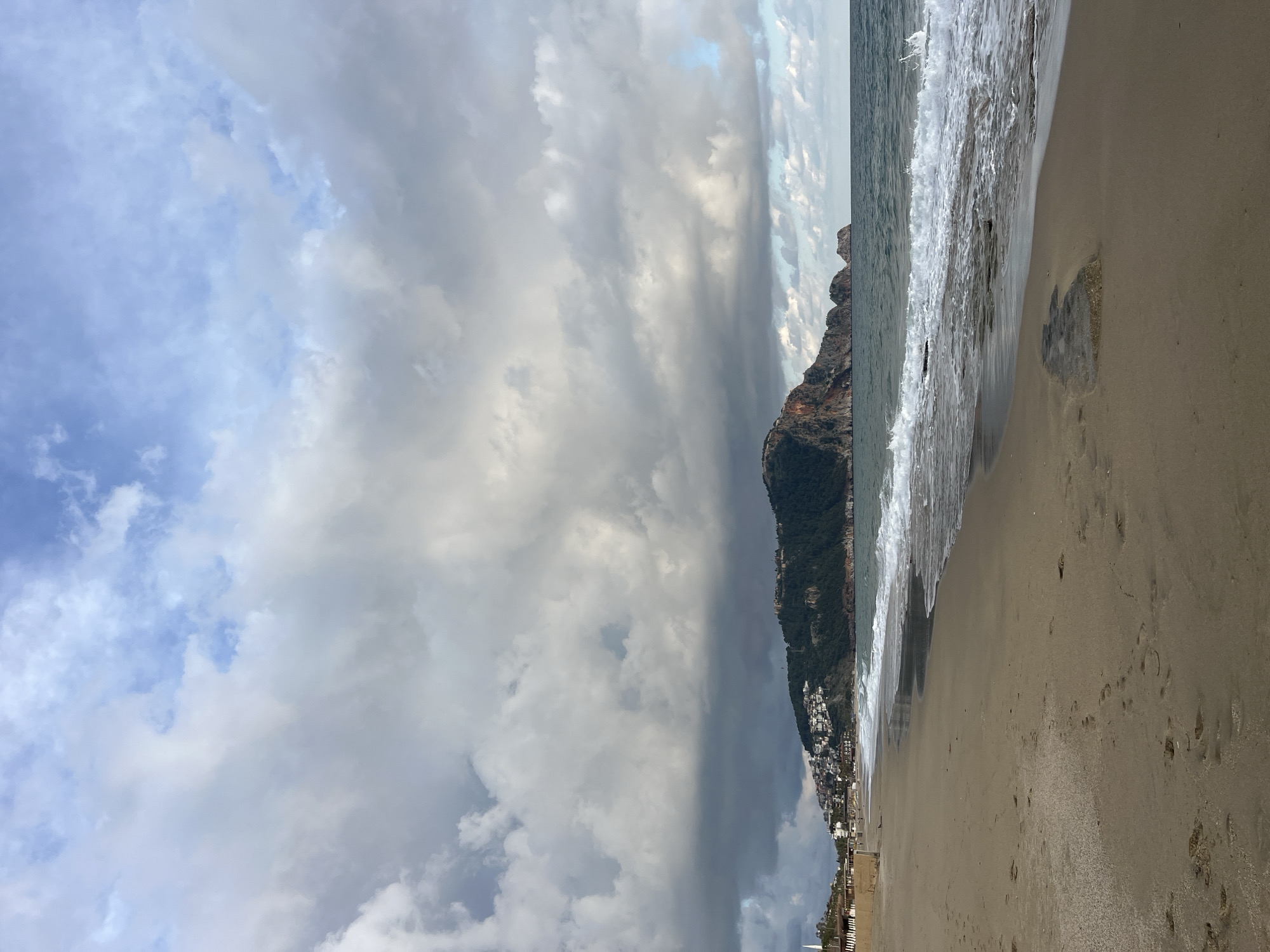 The image captures a beach scene in Alanya, Turkey. The photograph is taken from the shoreline, showcasing a sandy beach meeting the ocean's edge. Waves are visible, crashing onto the shore. A large mountain rises steeply from the water's edge and runs to the right of the image, partially covered in green vegetation, houses and buildings that appear to be part of the city. The sky is overcast with numerous gray and white clouds, creating a dramatic effect. Footprints can be seen in the sand, suggesting recent human presence. The scene presents a contrast between the natural elements of the sea, beach, and sky, and the built environment.