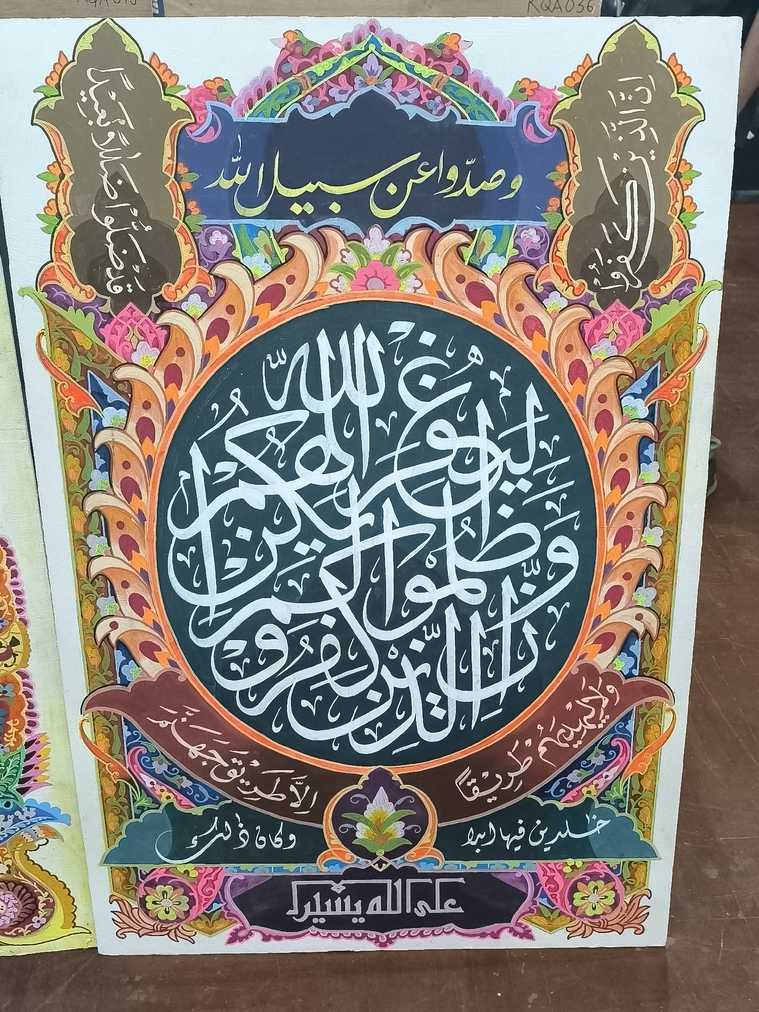 A vertical, eye-level, medium shot captures a colorful Islamic calligraphy artwork displayed against a white background. The artwork is framed with ornate patterns of flowers and leaves in vibrant hues of pink, orange, green, blue, and purple.
The central element is a large, circular motif filled with white Arabic script on a dark green background. This script is intricate and elaborate, typical of Islamic calligraphy. Surrounding the circle is a detailed border adorned with decorative elements and more floral patterns.
Above the circle, a dark blue banner features yellow Arabic script. To the left and right of this banner are two dark brown, shield-shaped panels, each containing white Arabic calligraphy.
Below the central circle, a dark brown curved banner displays white Arabic script. Underneath this, a dark green curved shape also contains white Arabic script.
At the very bottom of the artwork, a purple banner with white decorative elements hosts black Arabic script. The entire piece is vibrant and richly decorated, showcasing a complex arrangement of text and decorative art. The background appears to be a flat, light-colored surface, possibly a wall or a large board, and a portion of another similar artwork is visible to the left, suggesting a display or exhibition.
The scene is set in Minasa Te'ne, Indonesia, though the specific location within the city is not discernible from the image itself.
